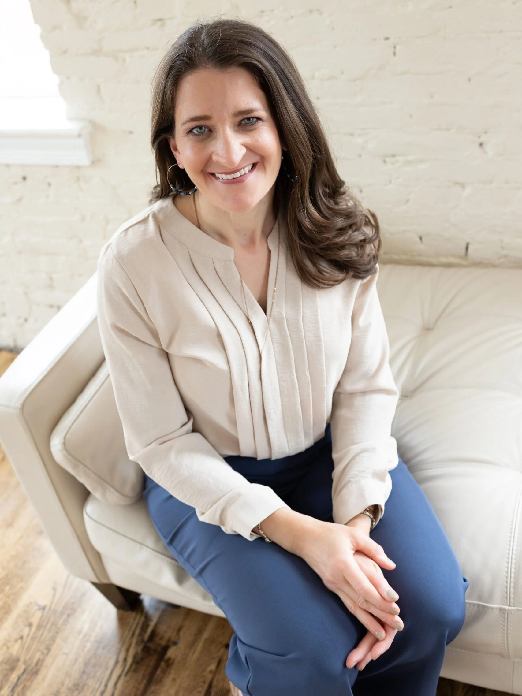 Kiersten Lortz with brown hair, blue eyes, and light skin, sitting on a beige sofa, smiling at the camera. She is wearing a beige blouse and blue pants, with a bracelet and hoop earrings, in a room with a white brick wall and wooden floor.