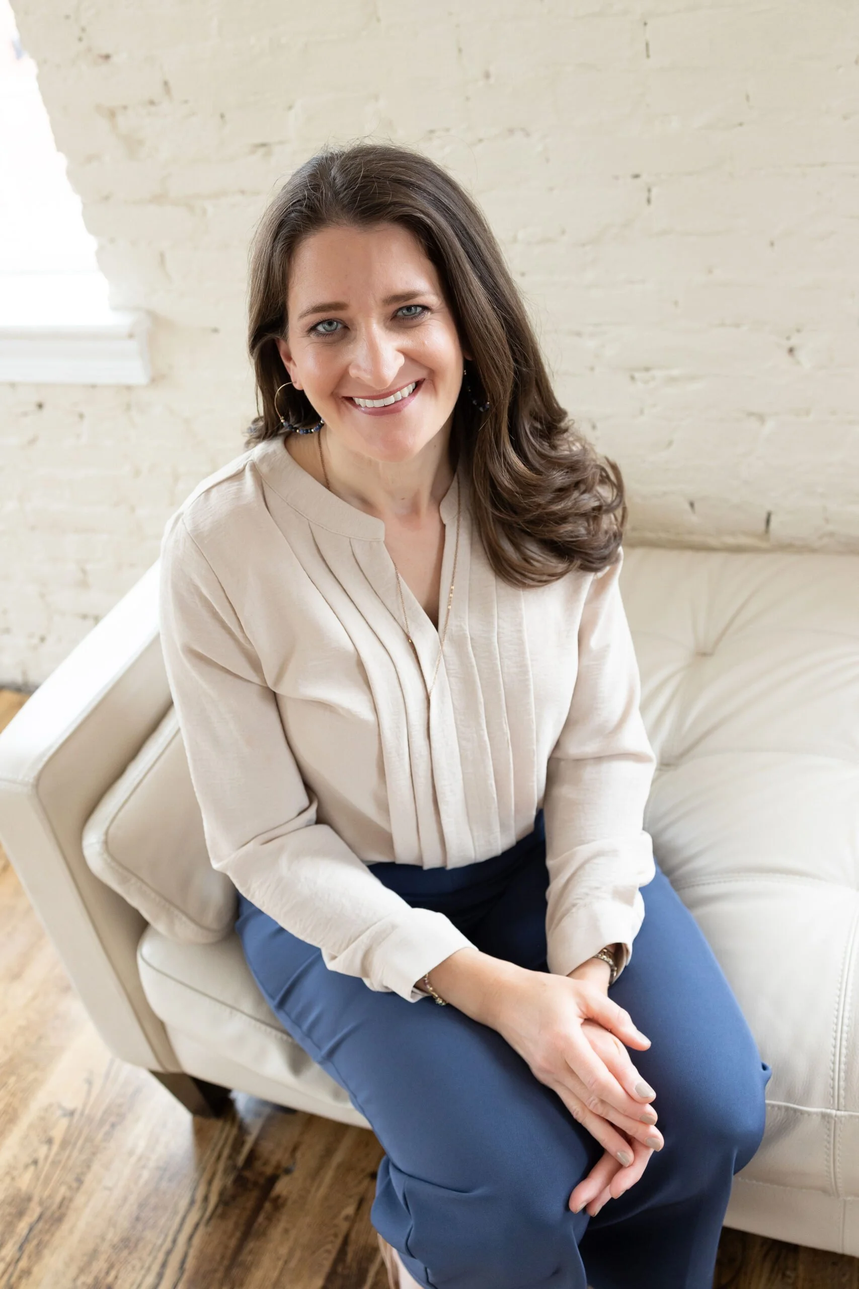A smiling Kiersten Lortz with long dark hair, wearing a cream-colored blouse and dark blue pants, sitting on a light-colored sofa in a room with white brick walls and wooden floors.