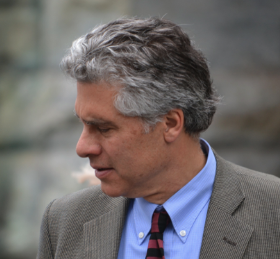 A man with gray hair in a gray blazer, blue shirt, and striped tie looking down outdoors.