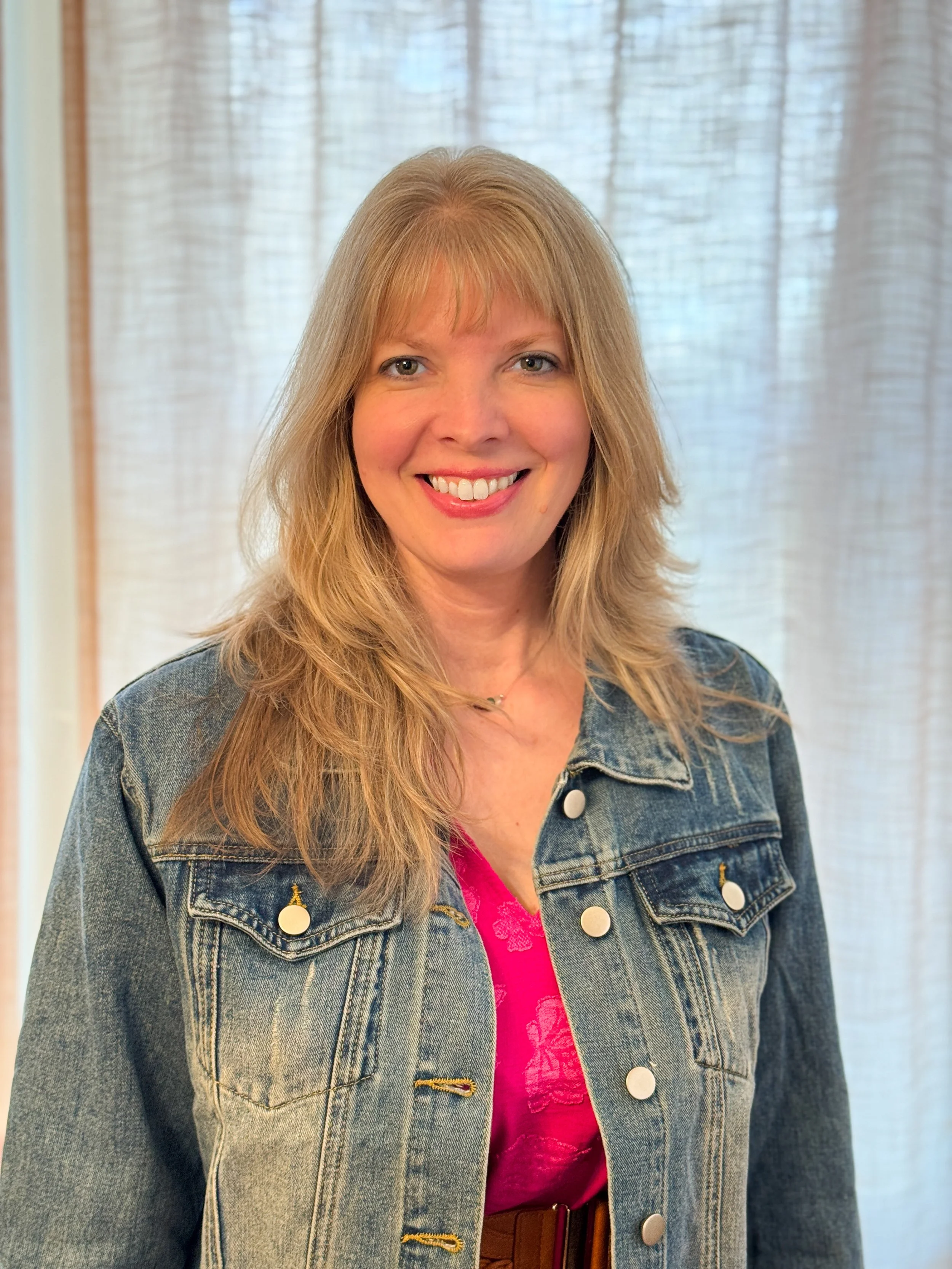 A woman with shoulder-length blonde hair, smiling, wearing a pink top and a denim jacket, standing in front of a light-colored curtain.