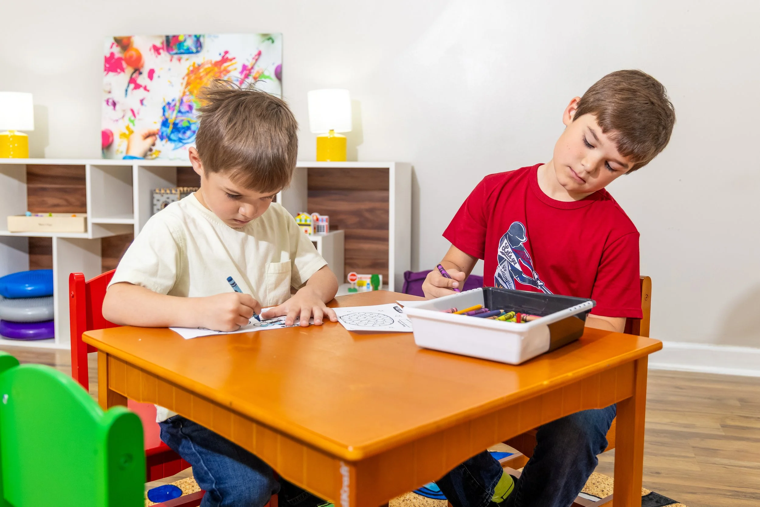 Two boys coloring with crayons during play therapy at a table in a classroom, with shelves and colorful artwork in the background