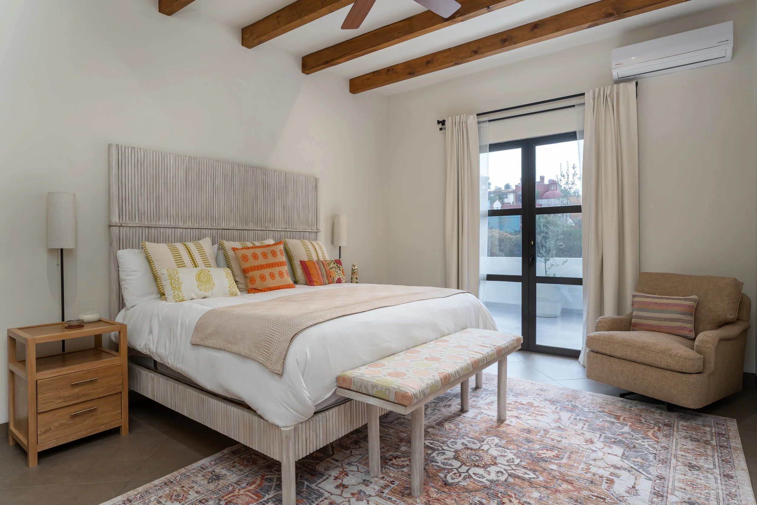 A bedroom with a large bed featuring colorful pillows, a wooden nightstand, a cushioned bench at the foot of the bed, a beige armchair with a striped pillow, and a sliding glass door with white curtains that leads to a balcony, with wooden ceiling beams and an air conditioning unit.