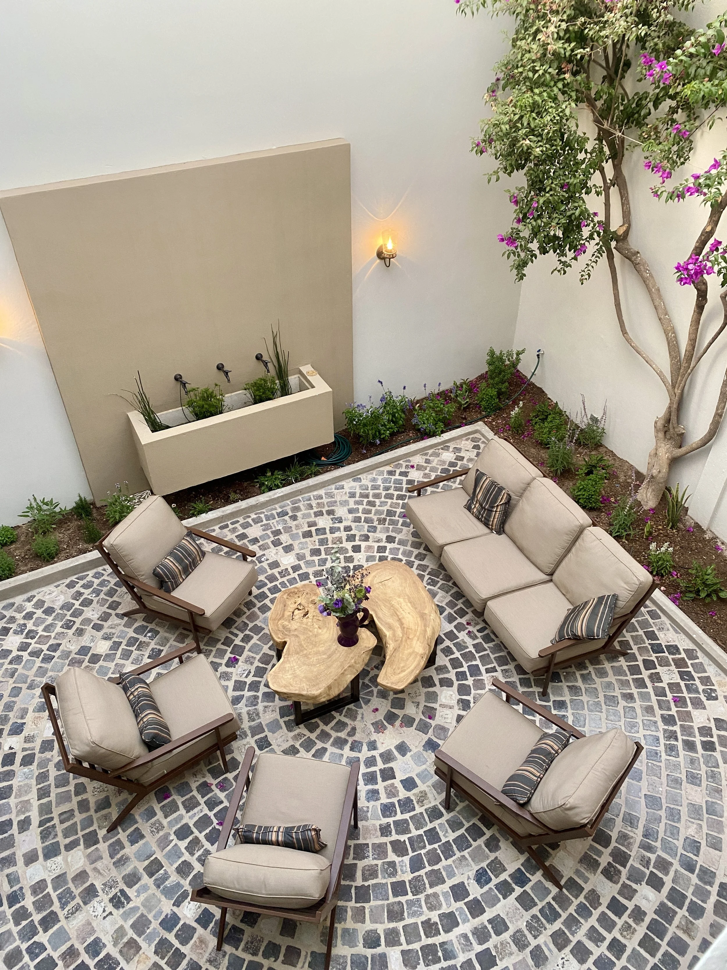 An outdoor patio with circular cobblestone flooring, beige cushioned chairs with striped pillows, a wooden coffee table with a flower vase, potted plants, and a bougainvillea tree against white walls.