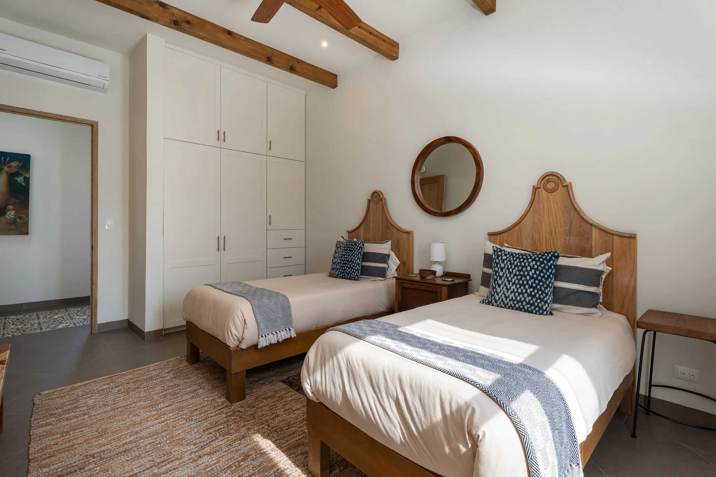 A bedroom with two twin beds with wooden headboards, a nightstand with a lamp between the beds, a mirror on the wall above the nightstand, and a white wardrobe in the corner. The room has a wooden beam ceiling, a rug on the floor, and a window allowing sunlight to enter.