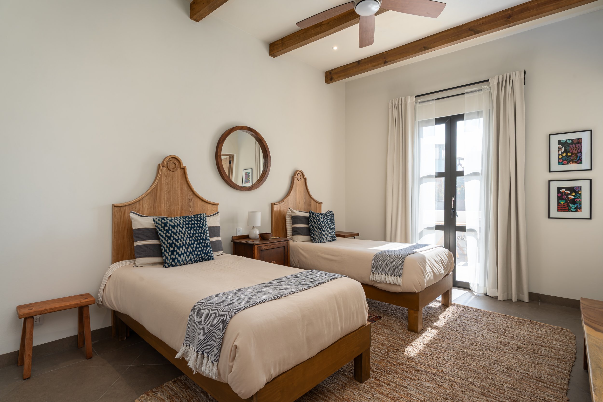 A bedroom with two single beds with wooden headboards and cream bedding, a nightstand with a lamp, a small wooden bench at the foot of one bed, a window with sheer white curtains, a ceiling fan, and framed artwork on the wall.