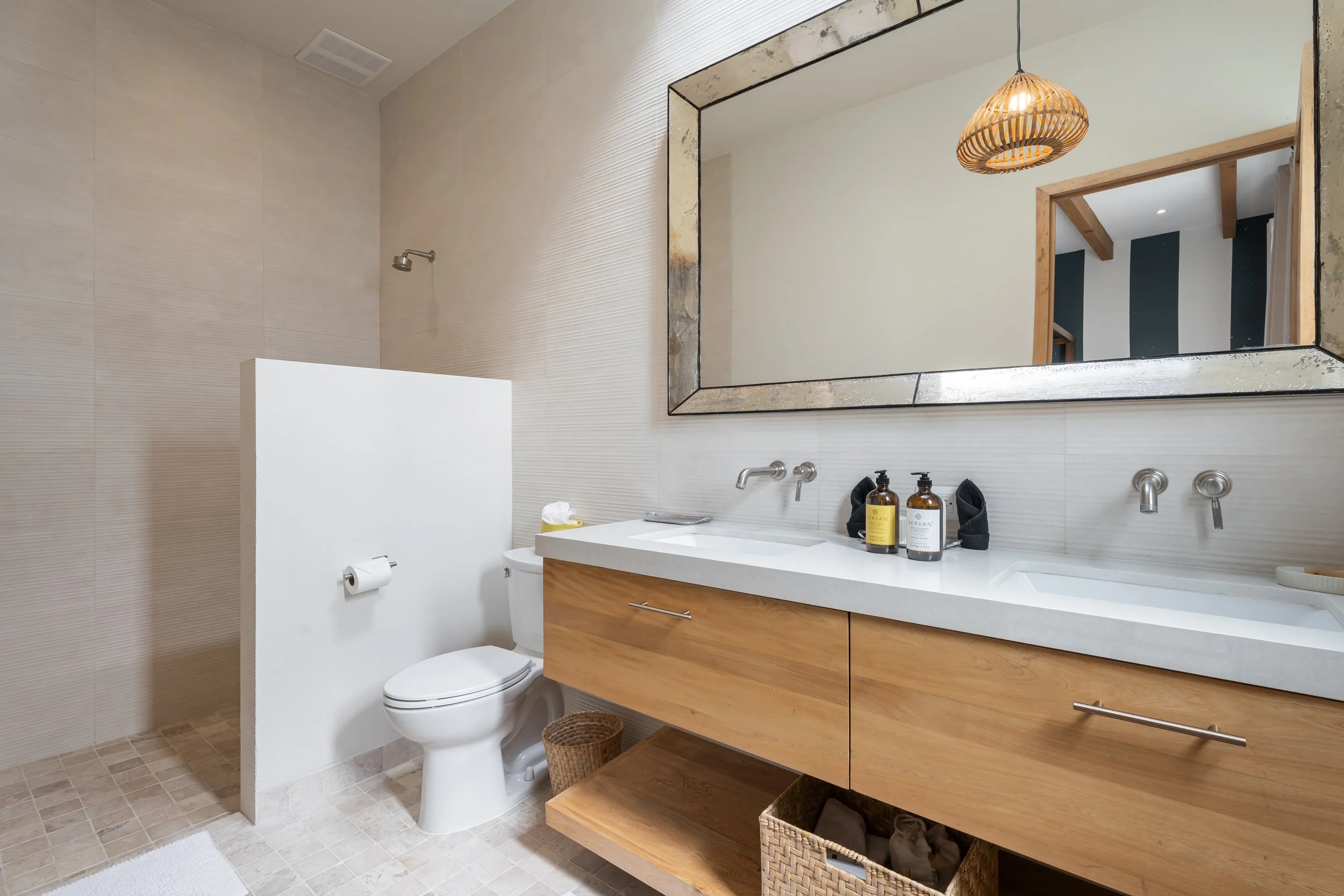 Modern bathroom with a large mirror, wooden vanity with a white countertop, and a toilet. The shower area is visible in the corner with a showerhead, and the bathroom has beige tiled walls and floors. Decorative lighting fixture is ceiling-mounted.