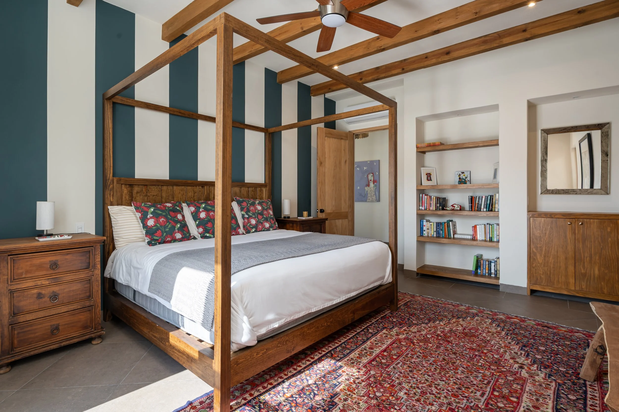 A bedroom with a wooden four-poster bed, a nightstand, and a bookshelf, featuring teal and white striped accent wall, wooden beams on ceiling, and a Persian rug.