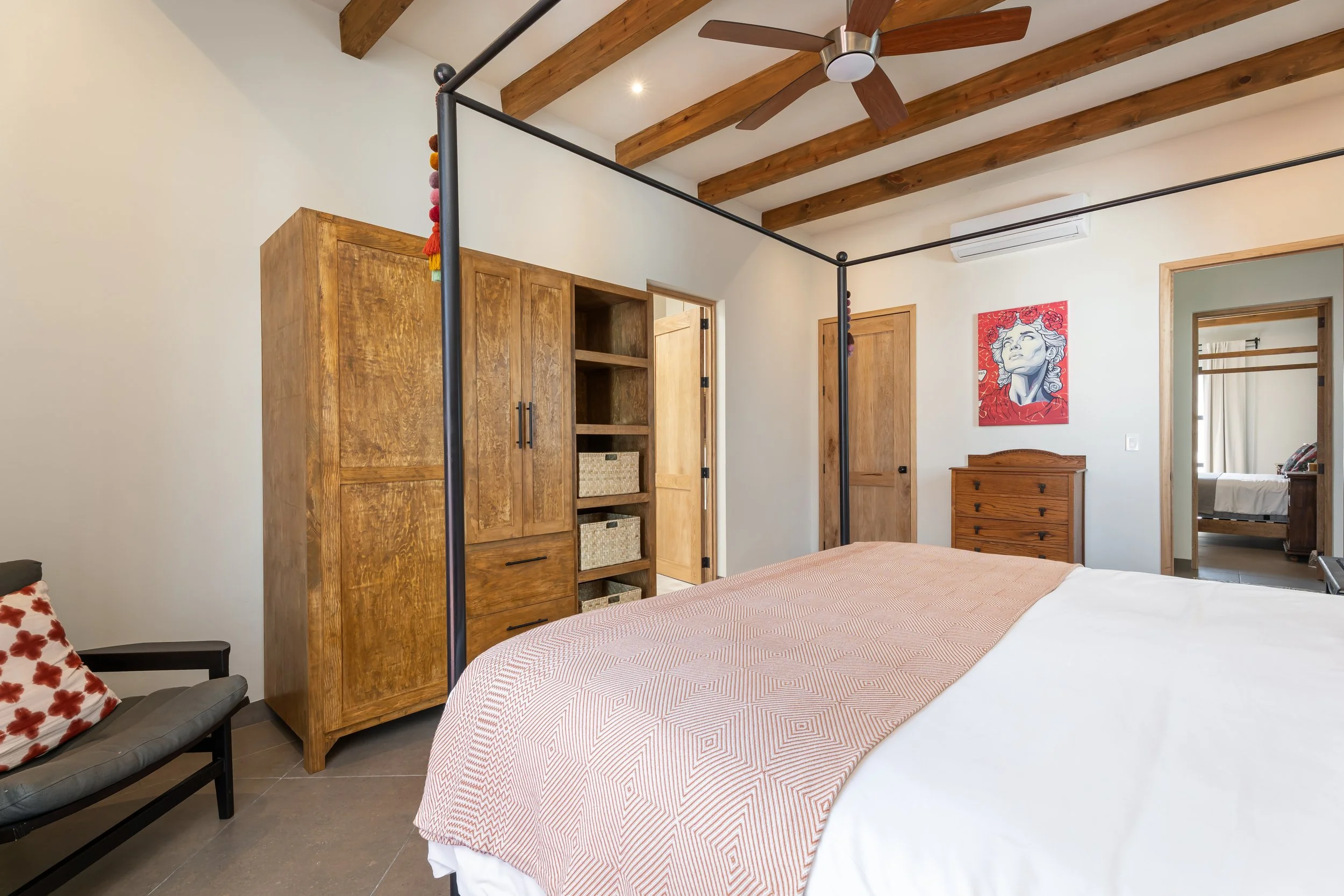 A bedroom with a wooden four-poster bed, nightstand, wood cabinets, and a colorful modern art portrait on the wall, ceiling beams, a ceiling fan, and an adjacent room visible through an open door.