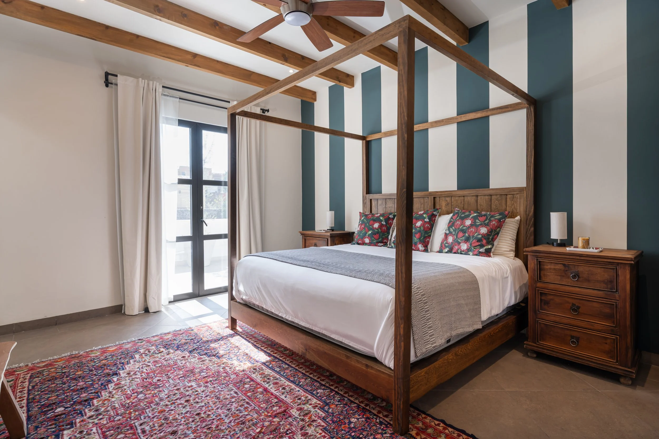 A bedroom with a large bed, wooden canopy frame, dark green and white striped accent wall, wooden nightstands, lamps, and a colorful oriental rug.