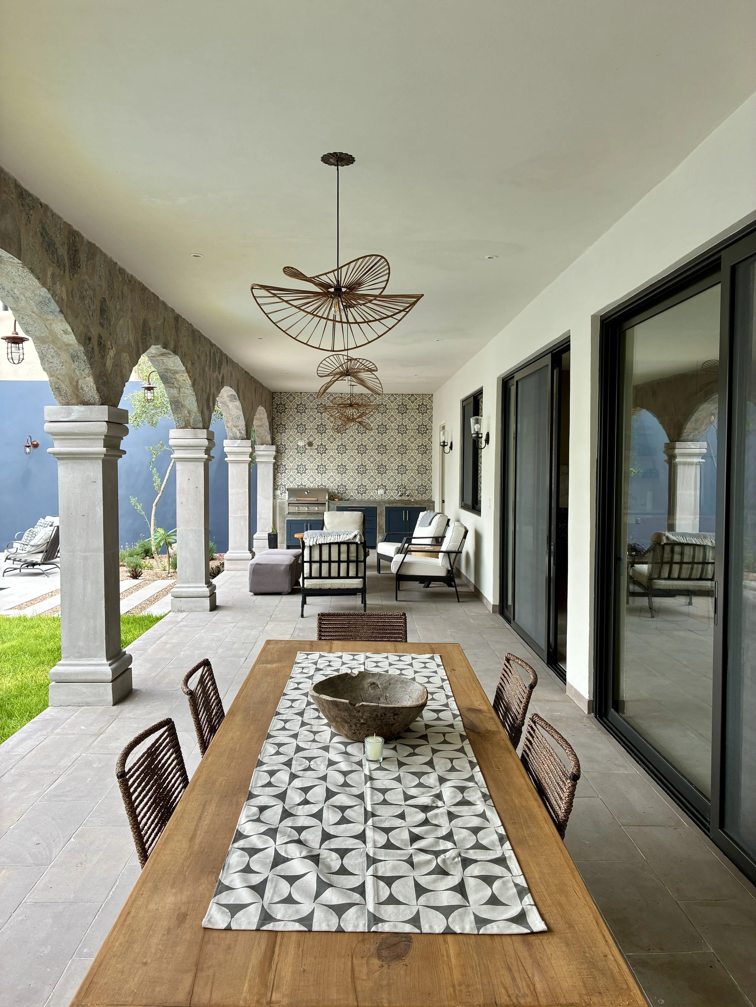Covered outdoor patio with a wooden dining table, six chairs, decorative bowl, and candle, featuring arched stone columns, modern seating area, patterned wall, and sliding glass doors.
