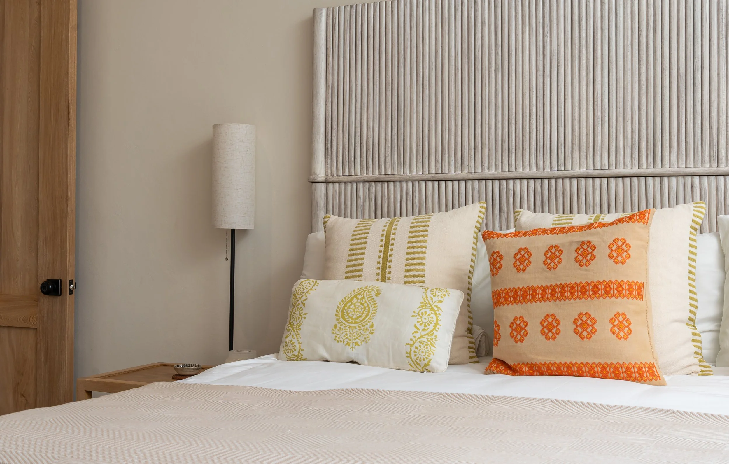 A neatly made bed with decorative pillows featuring orange, beige, and green patterns, a tall beige lamp beside a wooden nightstand, and a textured beige headboard in a bedroom.