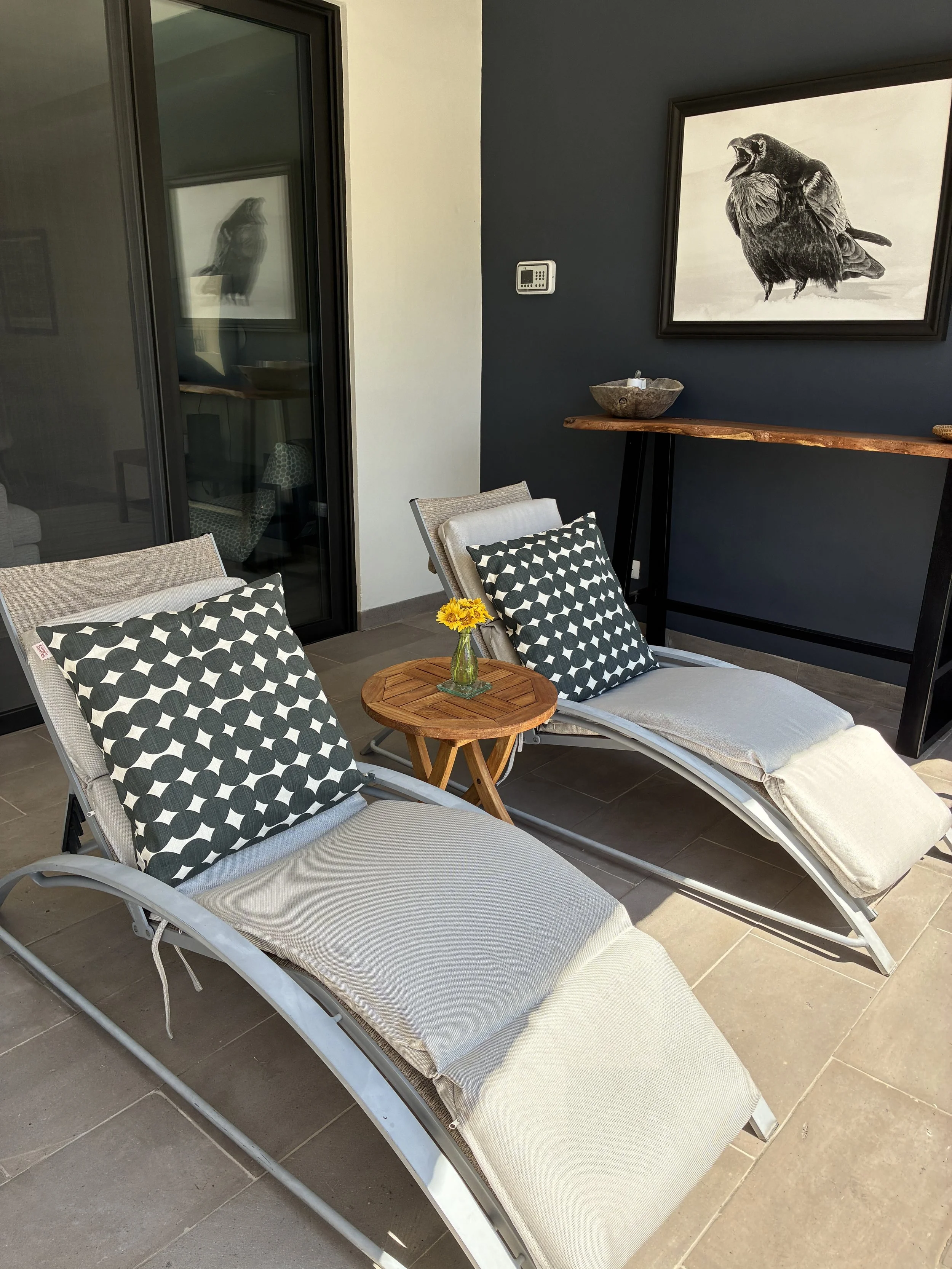 Two lounge chairs with black and white patterned pillows and beige cushions on a tiled patio, with a small wooden side table holding a yellow flower in a vase, against a gray and white wall with black framed artwork and a wooden console table.
