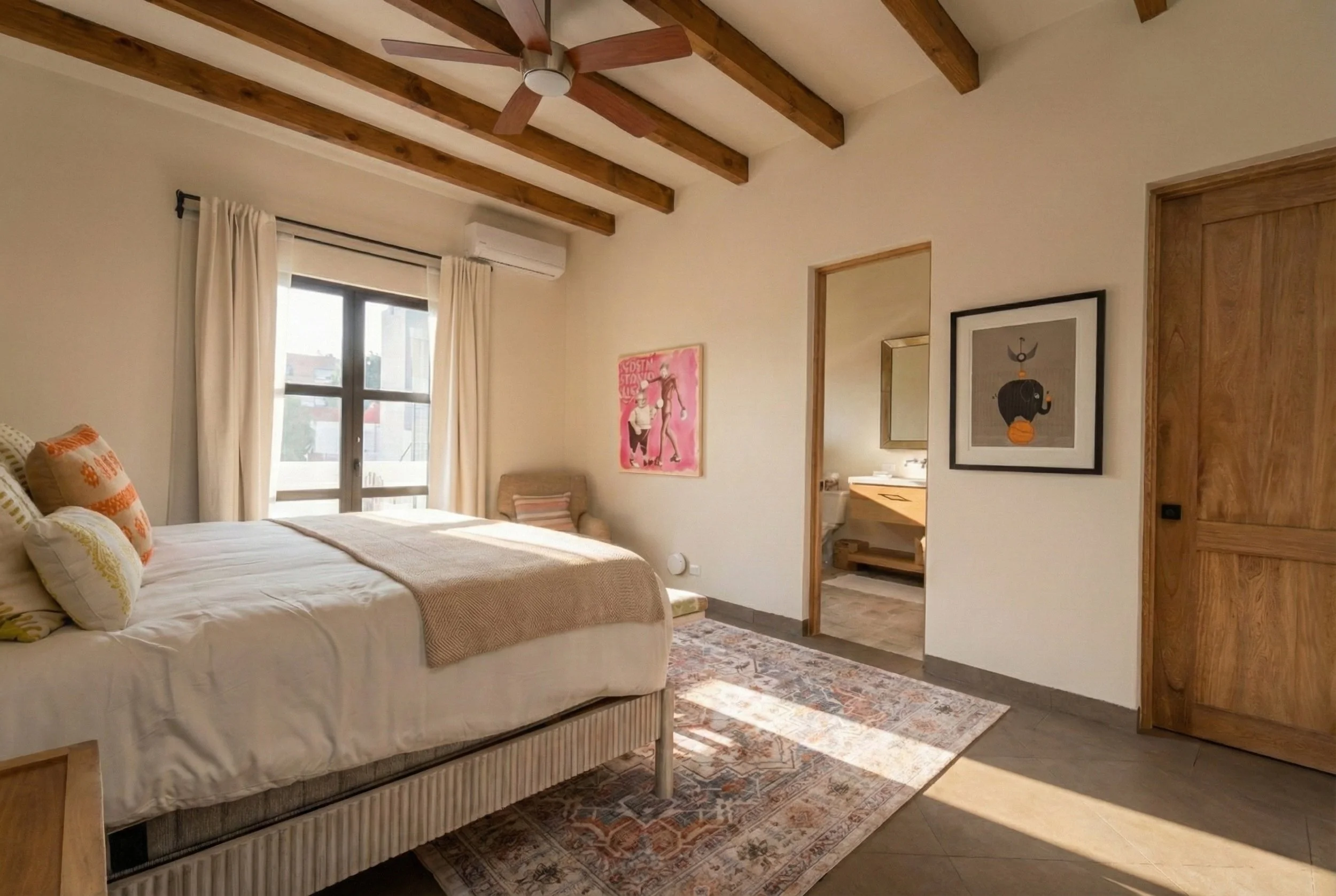 A cozy bedroom with a bed, decorative pillows, a chair, wall art, wooden ceiling beams, a window with curtains, and a view into a bathroom.