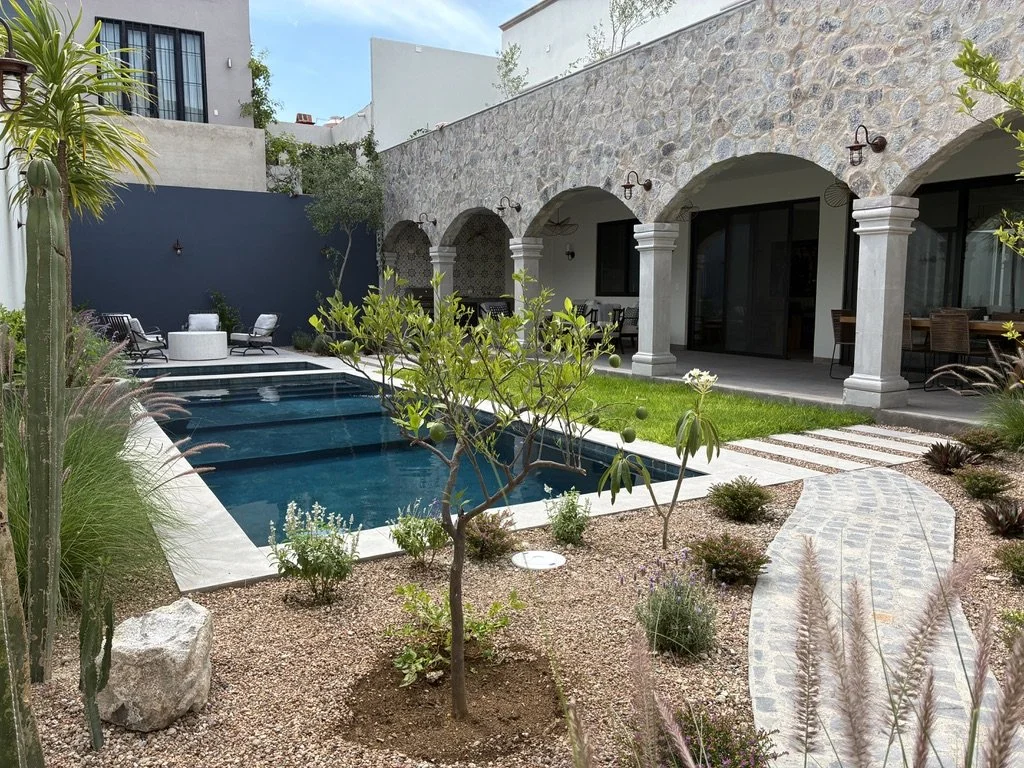 A modern backyard with a rectangular pool, lounging chairs, and a patio with arches supported by columns, all surrounded by desert-style landscaping with trees and plants.