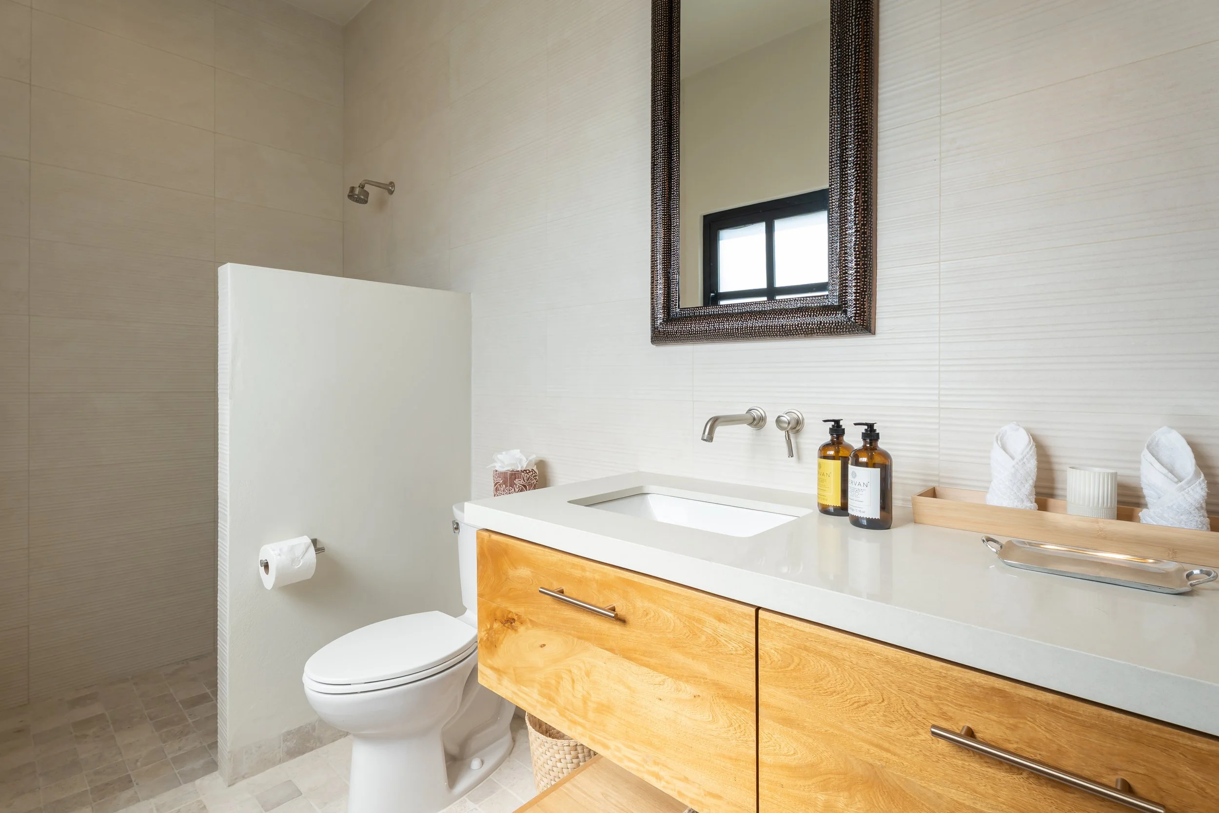 A clean bathroom with a white toilet, a wood vanity with a white countertop, a mirror, a window, and soap and towel accessories.