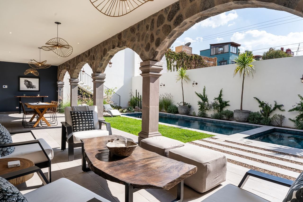 Indoor-outdoor living space with stone arches, modern furniture, and a swimming pool, overlooking neighboring buildings, with plants along the white wall.