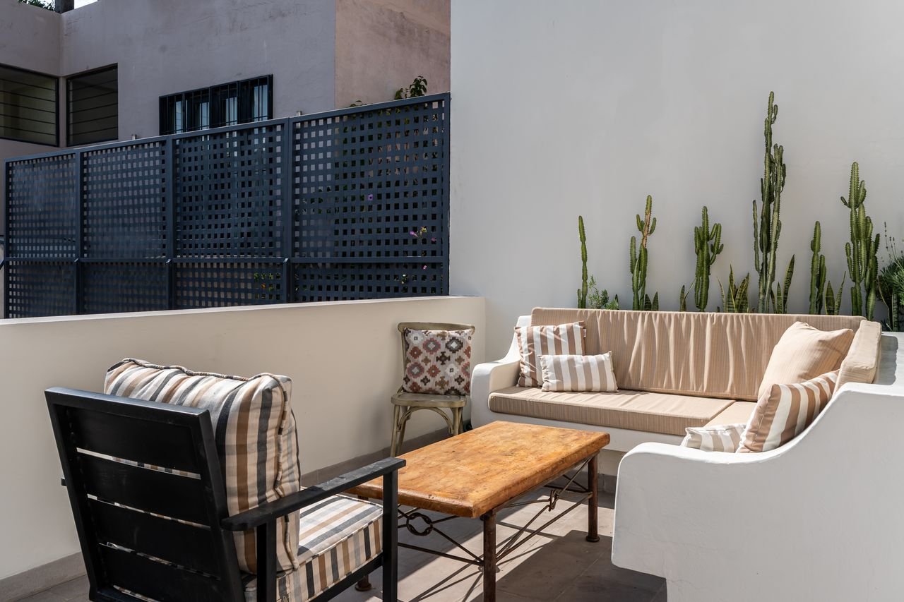 Outdoor patio seating with a beige sofa, striped throw pillows, a wooden coffee table, a black and beige armchair with striped cushions, a small decorative pillow on a wooden chair, tall cactus plants, a white wall, and a blue privacy fence.