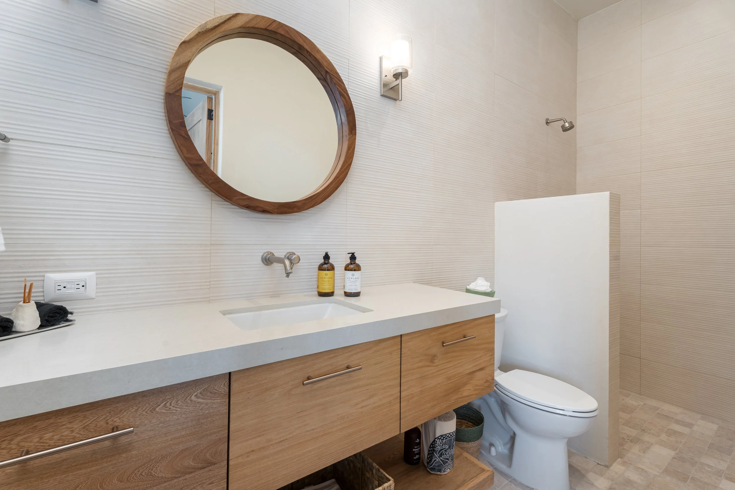 Modern bathroom with a wooden vanity, white countertop, round mirror, wall-mounted light, and a toilet. Bottles of soap or lotion are on the counter. Shower area is visible with a partial divider and tiled walls.