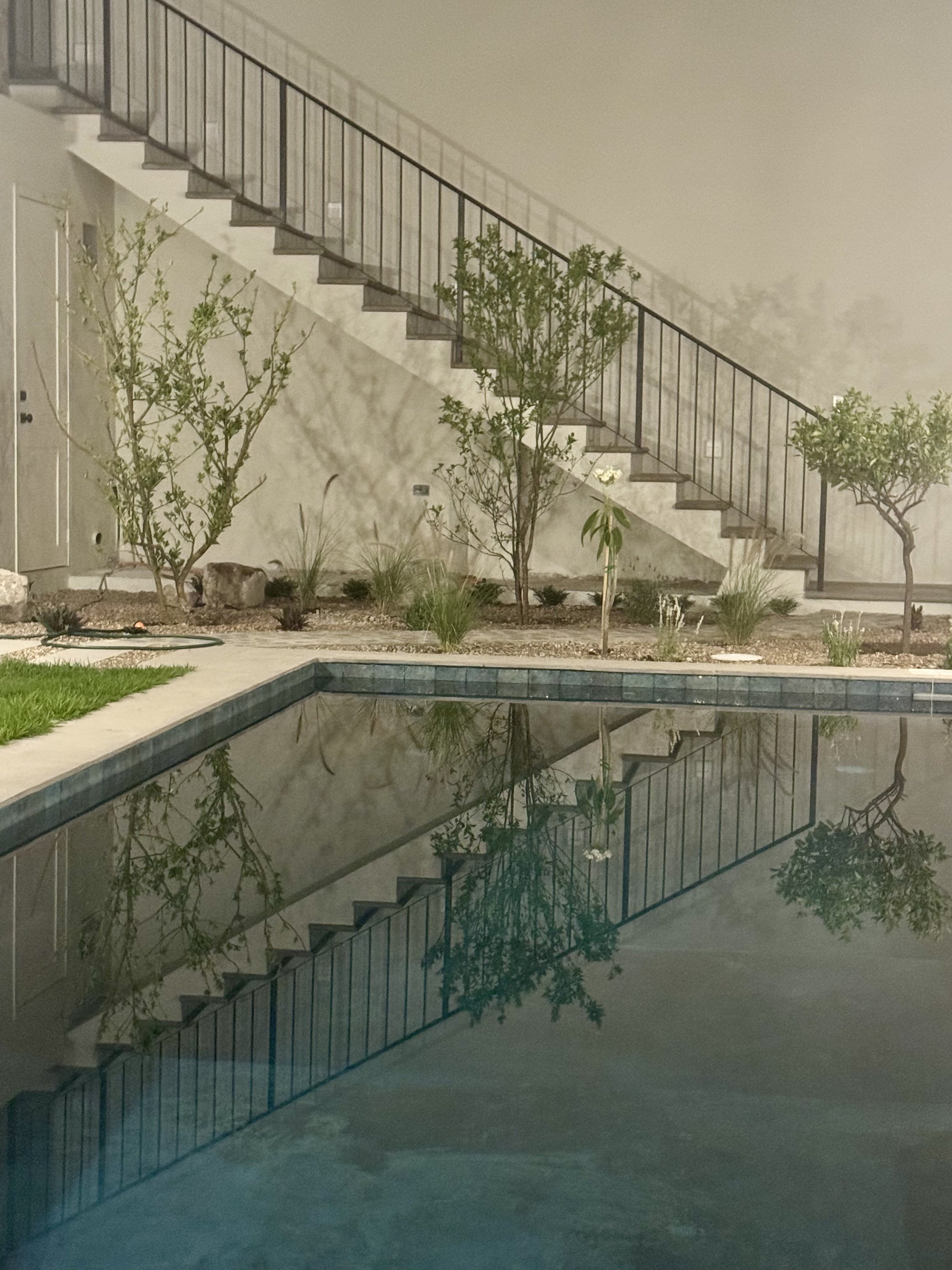 Nighttime backyard scene with a swimming pool reflecting the nearby trees, bushes, and staircase with a black metal railing.