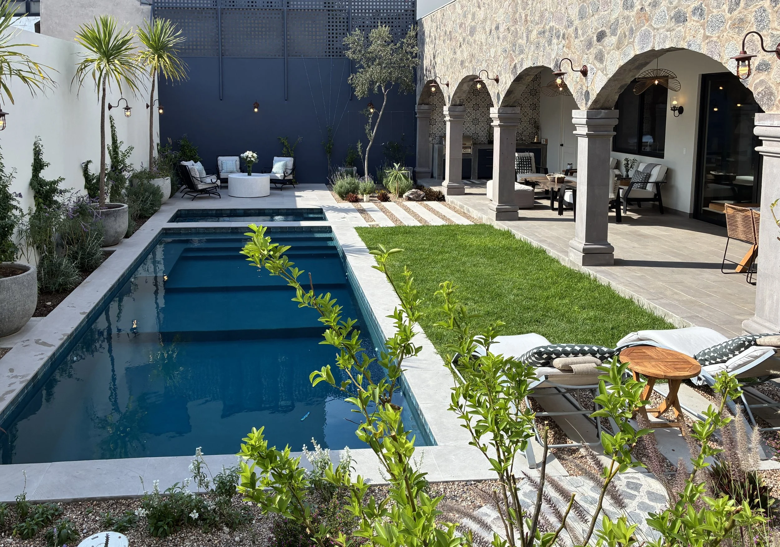 Backyard with swimming pool, sun loungers, small round table, garden plants, potted palms, shaded patio with seating area, stone archways, and outdoor lighting.