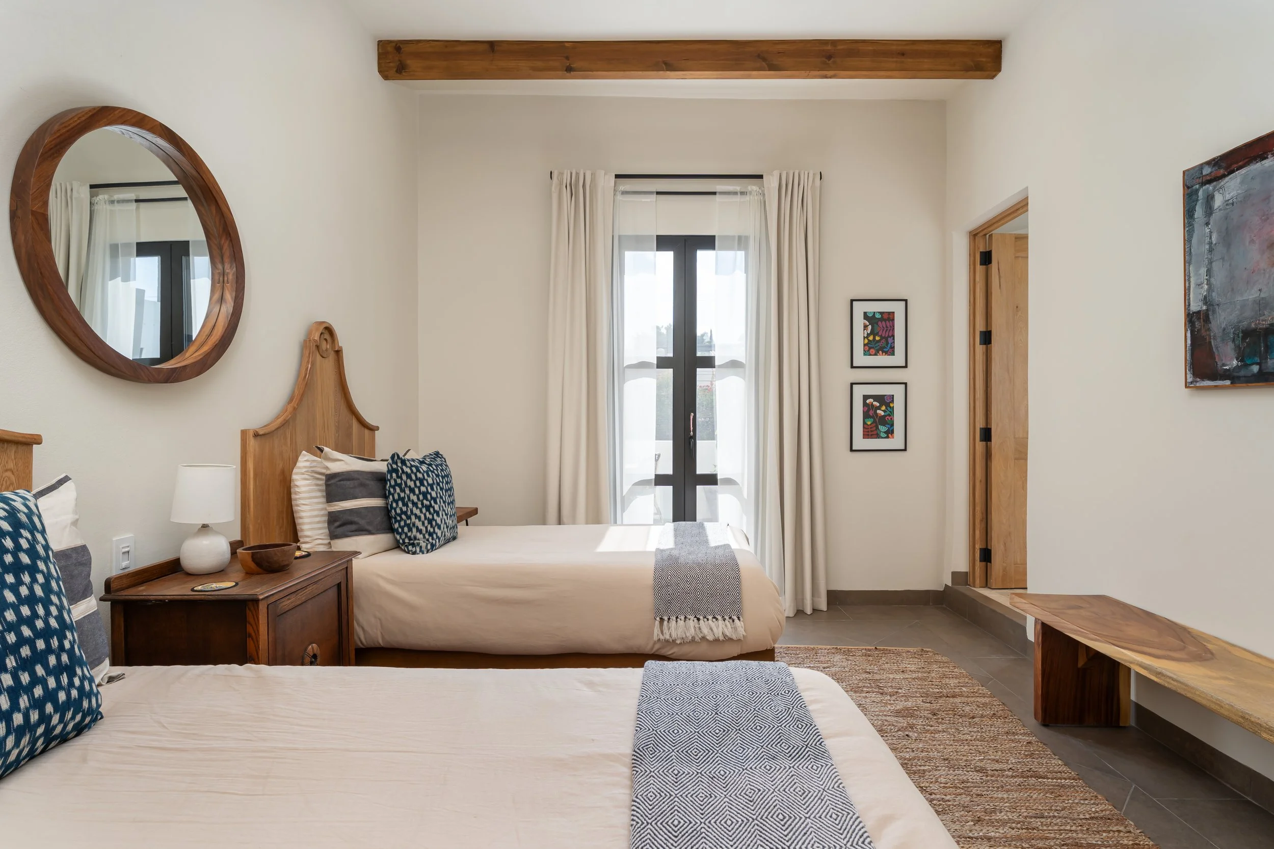 A bedroom with two single beds each with blue and white pillows, a wooden nightstand with a white lamp, a mirror on the wall, a window with white curtains, and framed artwork on the walls.