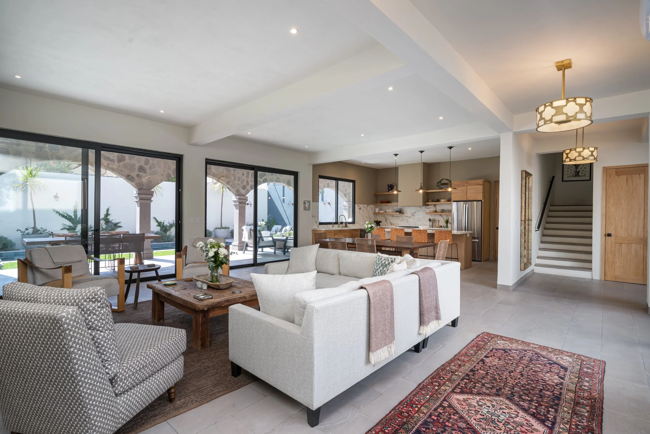 Open-concept living room with large glass sliding doors opening to an outdoor patio, featuring a white sectional sofa, a patterned armchair, a rustic wooden coffee table, and a kitchen with a marble island and open shelves.