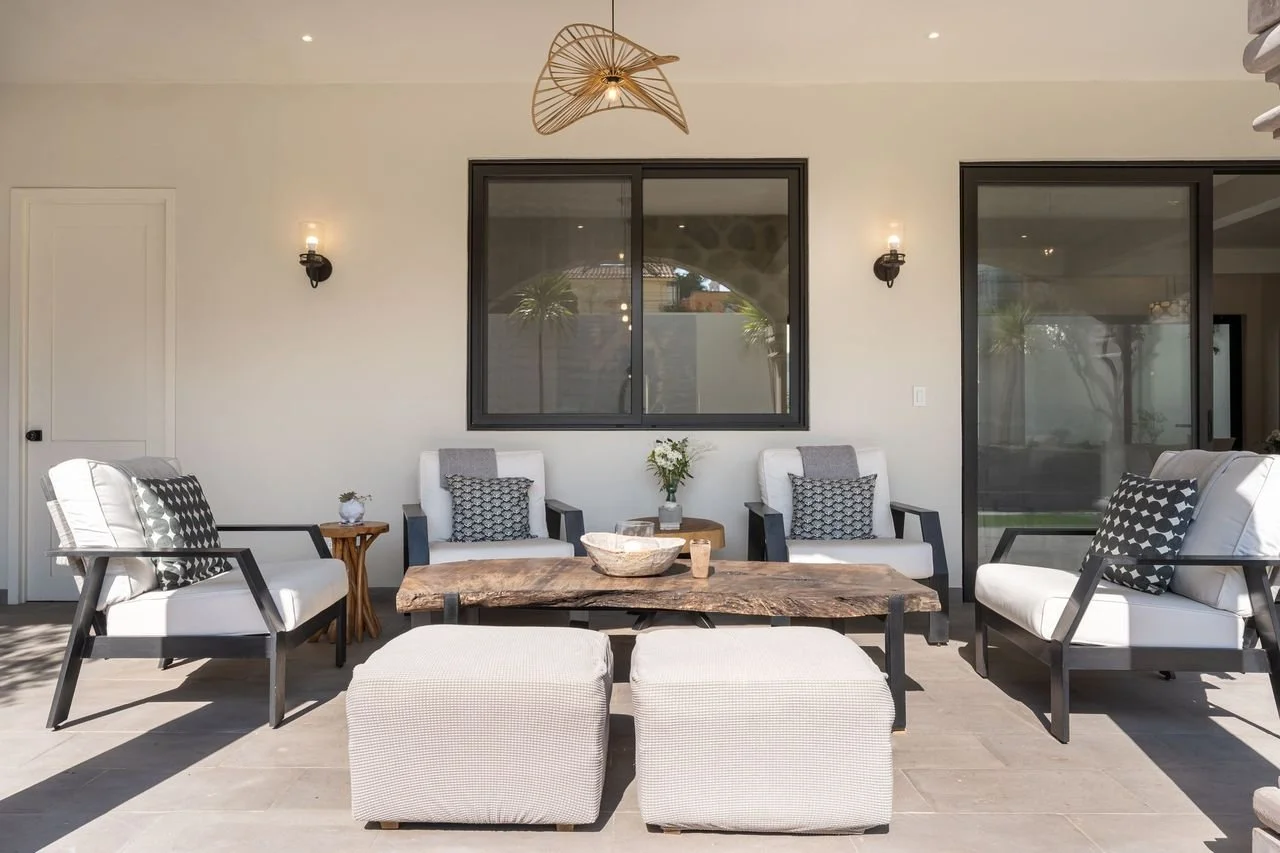 Outdoor patio with white and black seating, a rustic wooden coffee table, and decorative pillows, with a neutral wall and large windows in the background.