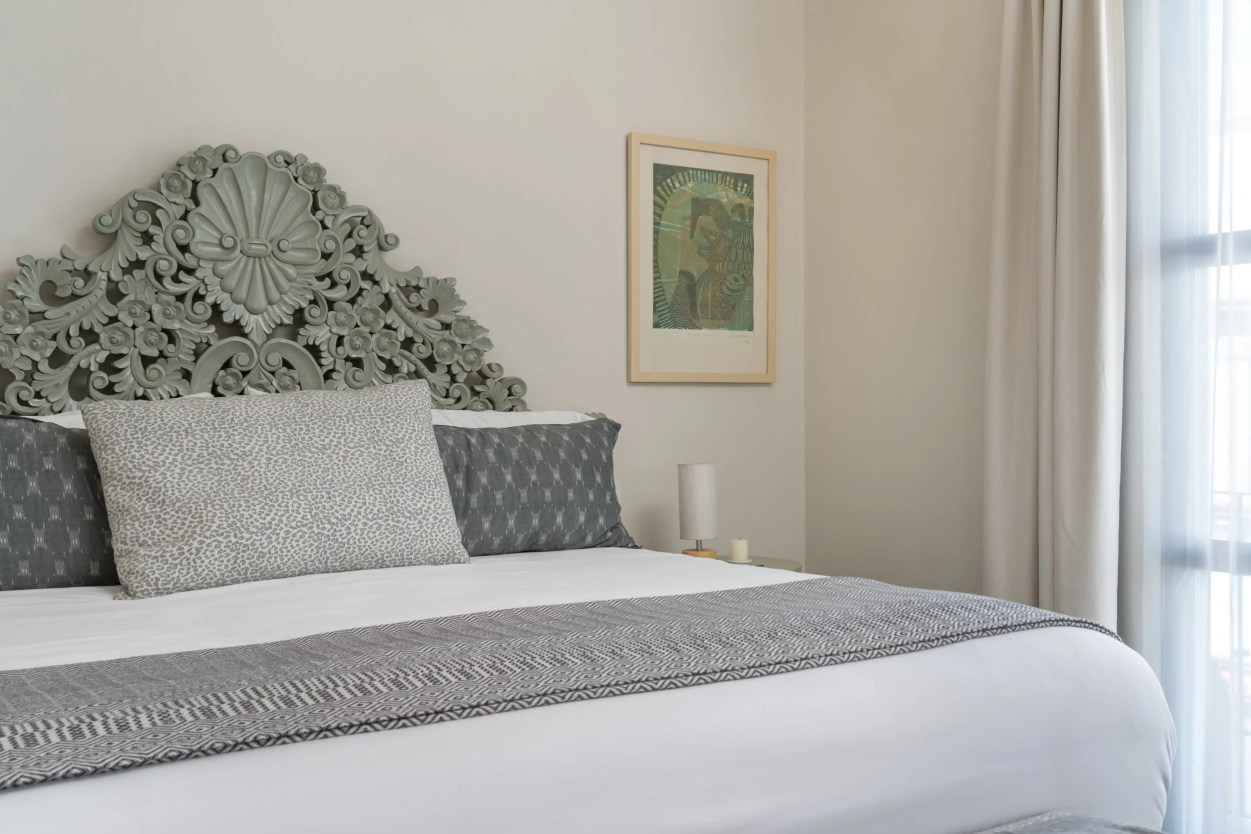 A neatly made bed with a decorative headboard, patterned pillows, a lamp, a small candle, and a framed artwork on a light-colored wall next to a window with white curtains.