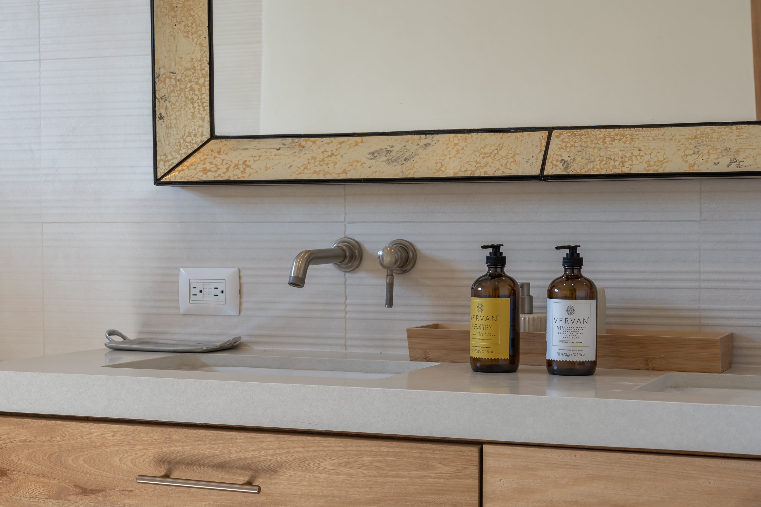 Bathroom sink with a mirror above, a wall outlet, and two bottles of liquid soap or lotion on a wooden tray.