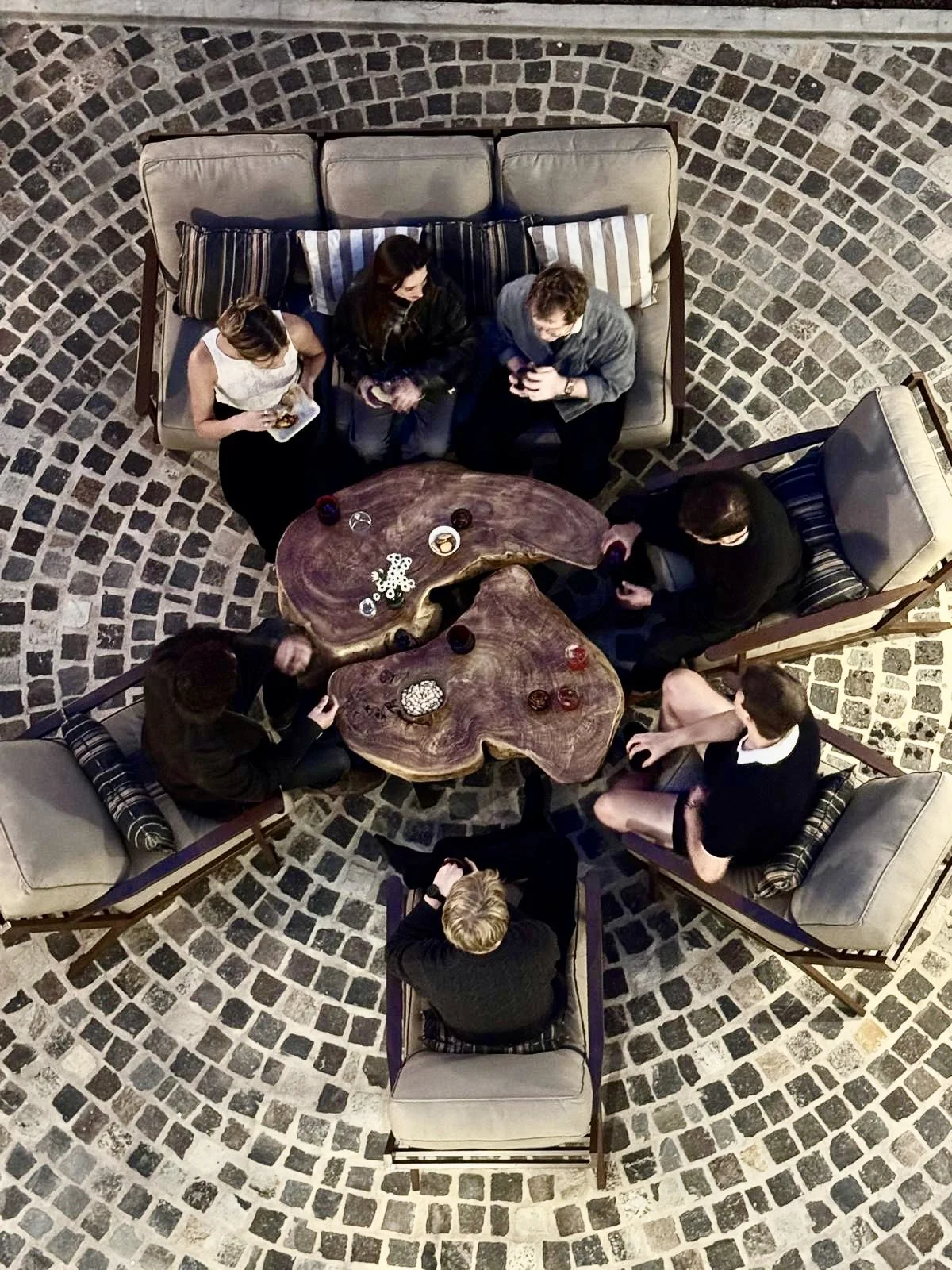 Aerial view of seven people gathered around a uniquely shaped wooden table and sitting on chairs and a sofa in a room with a circular stone-paved floor.