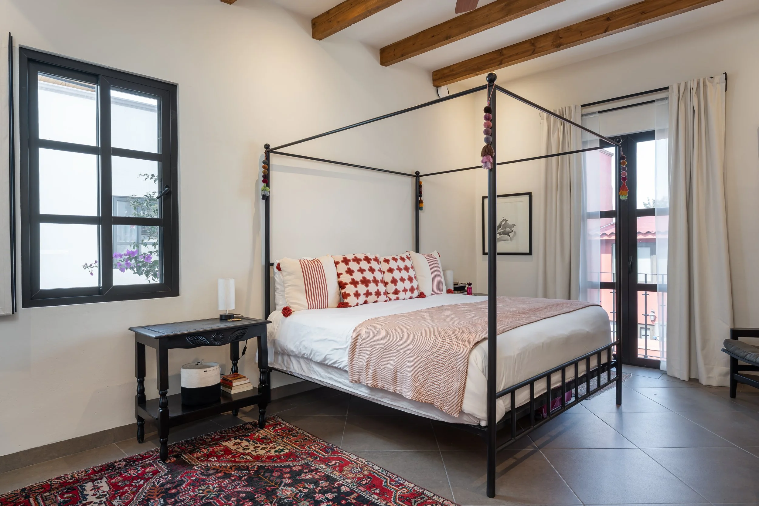 A bedroom with a black metal four-poster bed, white walls, wooden ceiling beams, two windows with black frames, and a balcony door. The bed has white bedding with pink and red pillows. There is a black nightstand with books, a lamp, and a small decorative object on it, and a patterned rug on the floor.