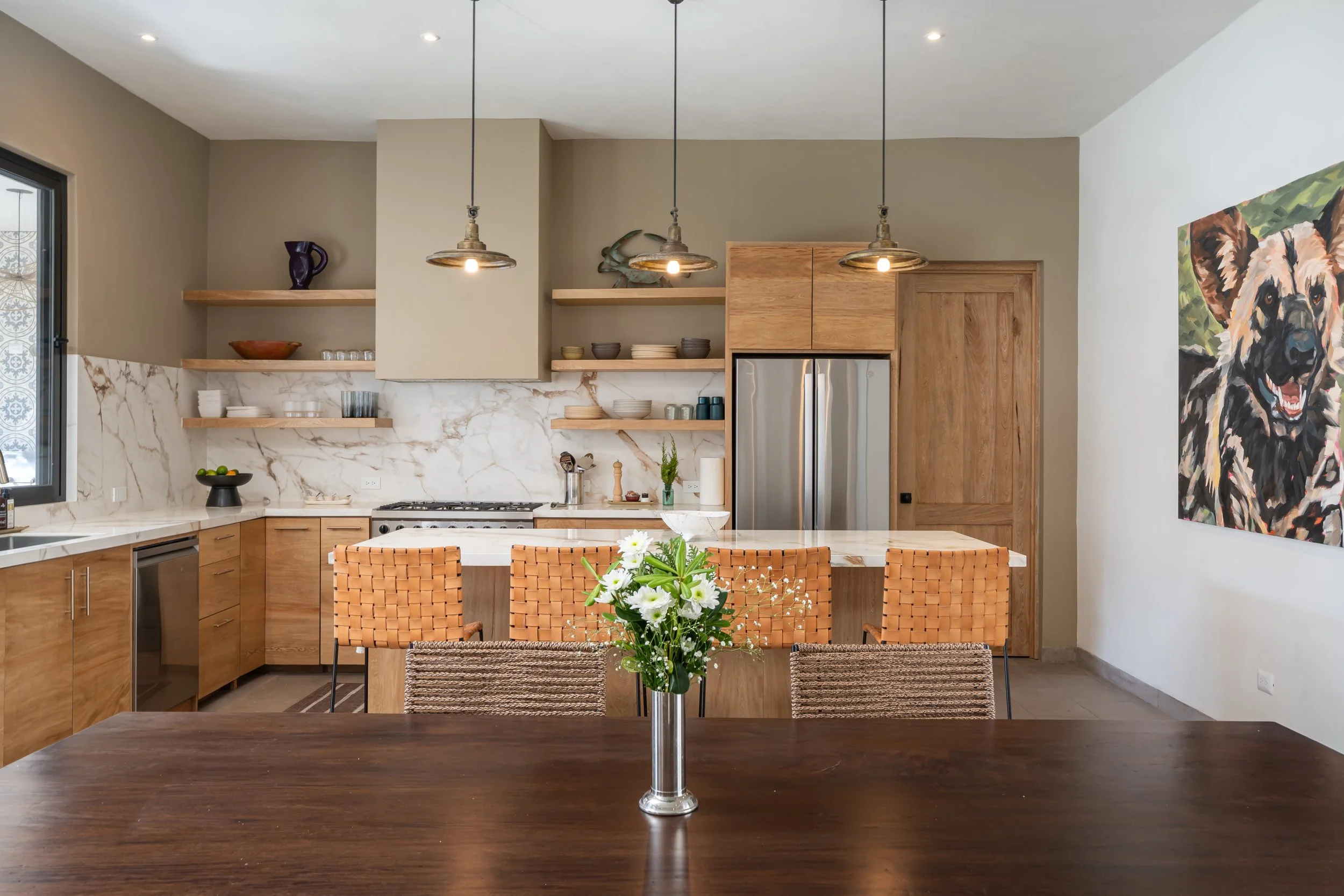Modern kitchen with marble backsplash, wooden shelves, stainless steel refrigerator, and a wooden dining table with a flower vase.