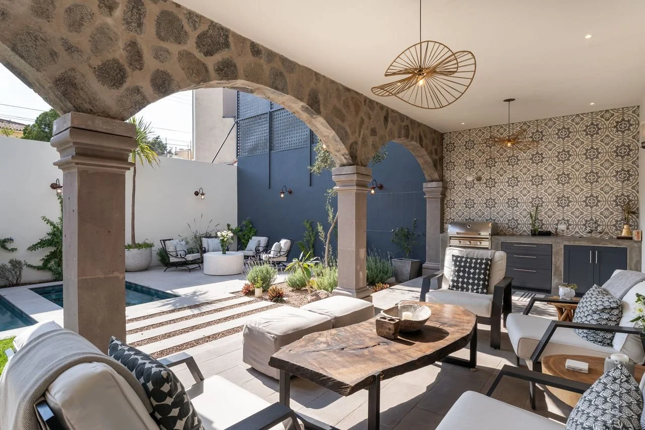 Covered outdoor patio with stone arched columns, a seating area with white and black patterned cushions, a wooden table, and an outdoor kitchen with patterned tile backsplash and gray cabinets. There is a small pool and various plants in the background.
