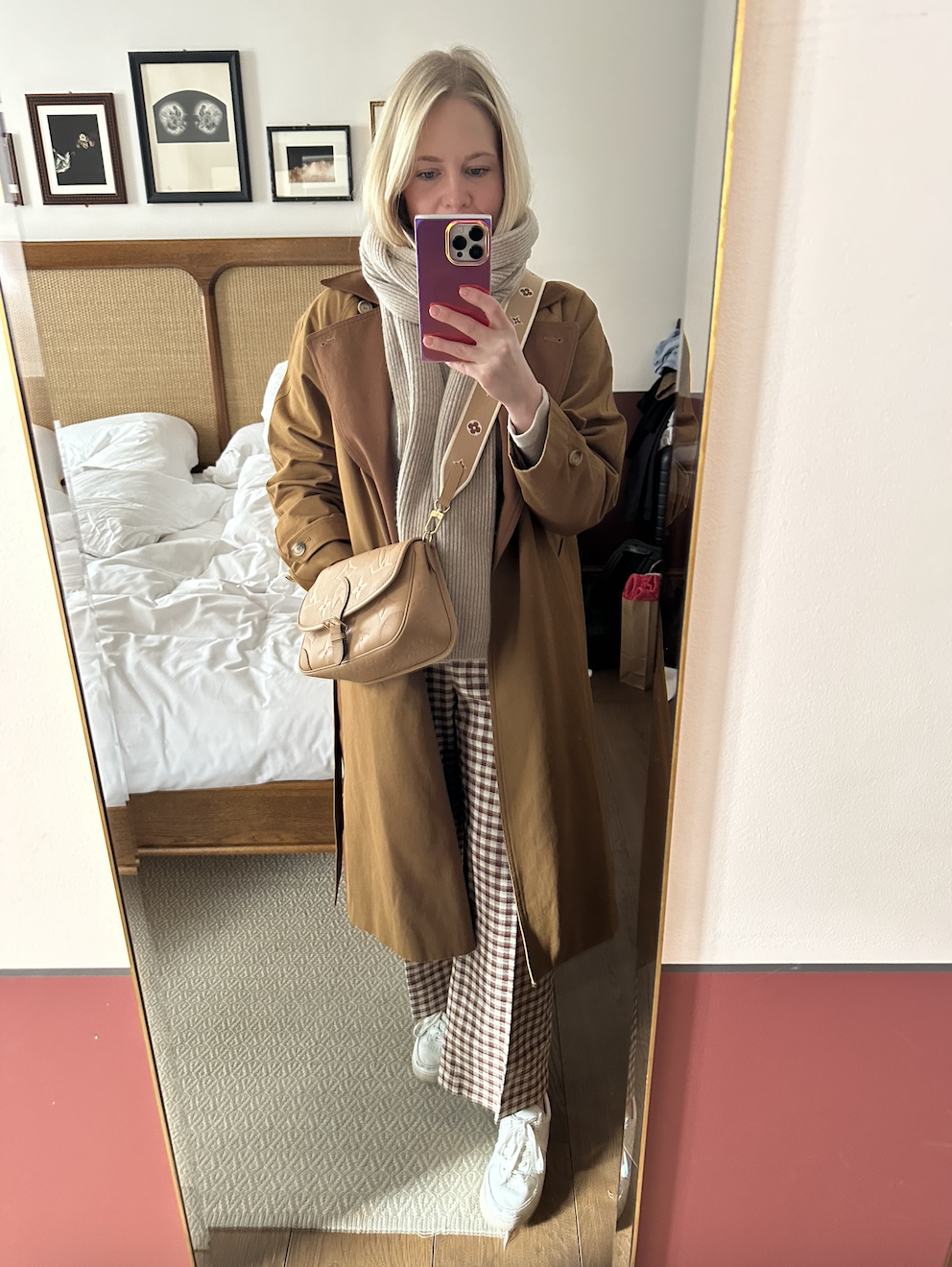 A woman taking a mirror selfie in a hotel room, wearing a brown trench coat, beige knit scarf, checked pants, white sneakers, and carrying a beige shoulder purse.