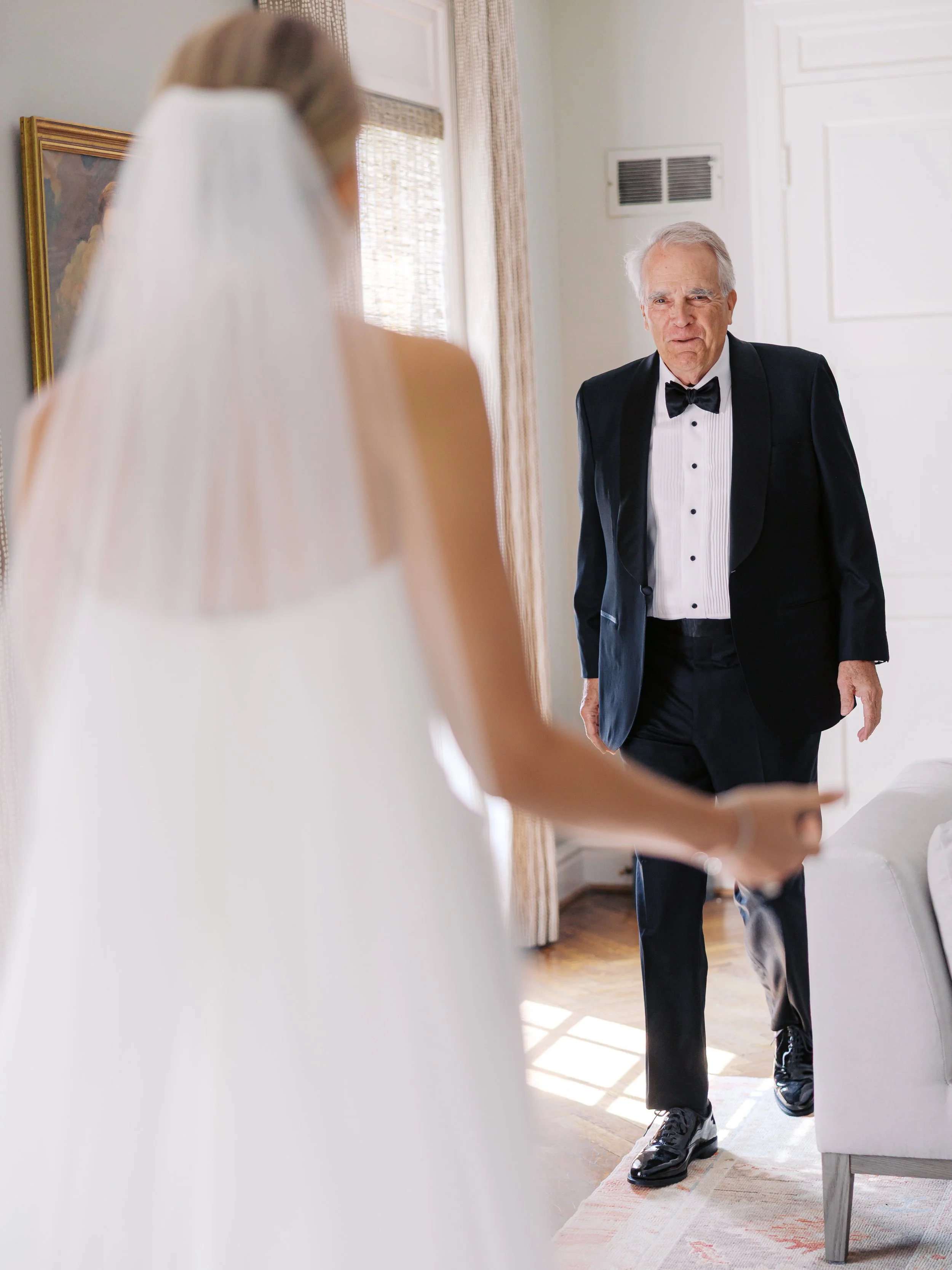 An older man in a tuxedo stands in a room, facing a woman in a wedding dress. The woman is out of focus and has her arm extended.