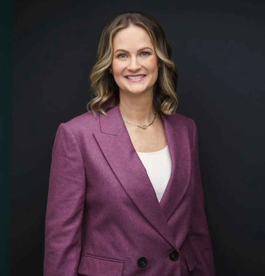 A woman with shoulder-length wavy blonde hair, wearing a purple blazer over a white top, smiling against a dark background.