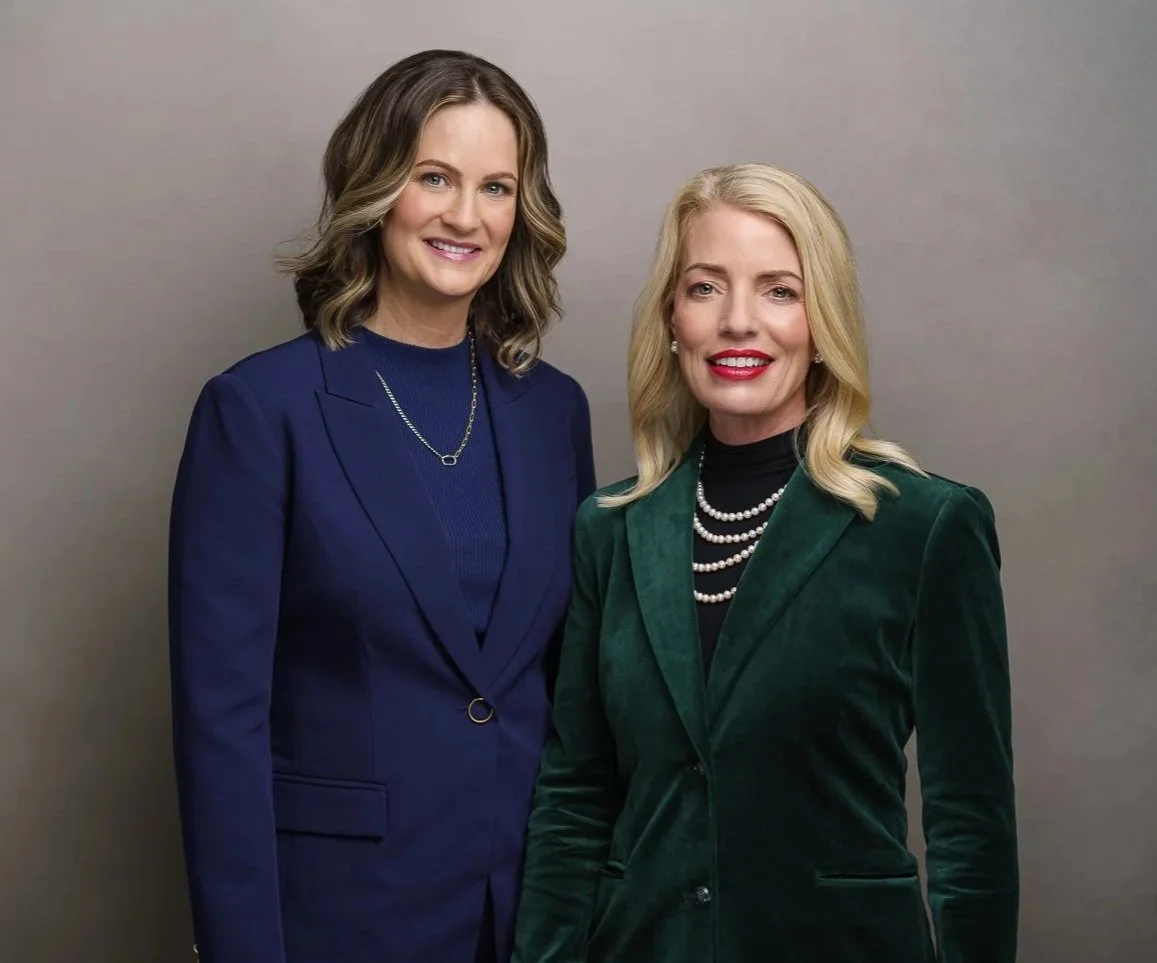 Two professional women standing side by side in front of a plain gray background, dressed in business attire and smiling at the camera.