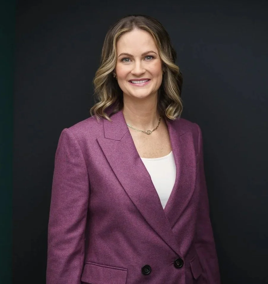 A woman in a purple blazer and white top standing against a dark background, smiling at the camera.