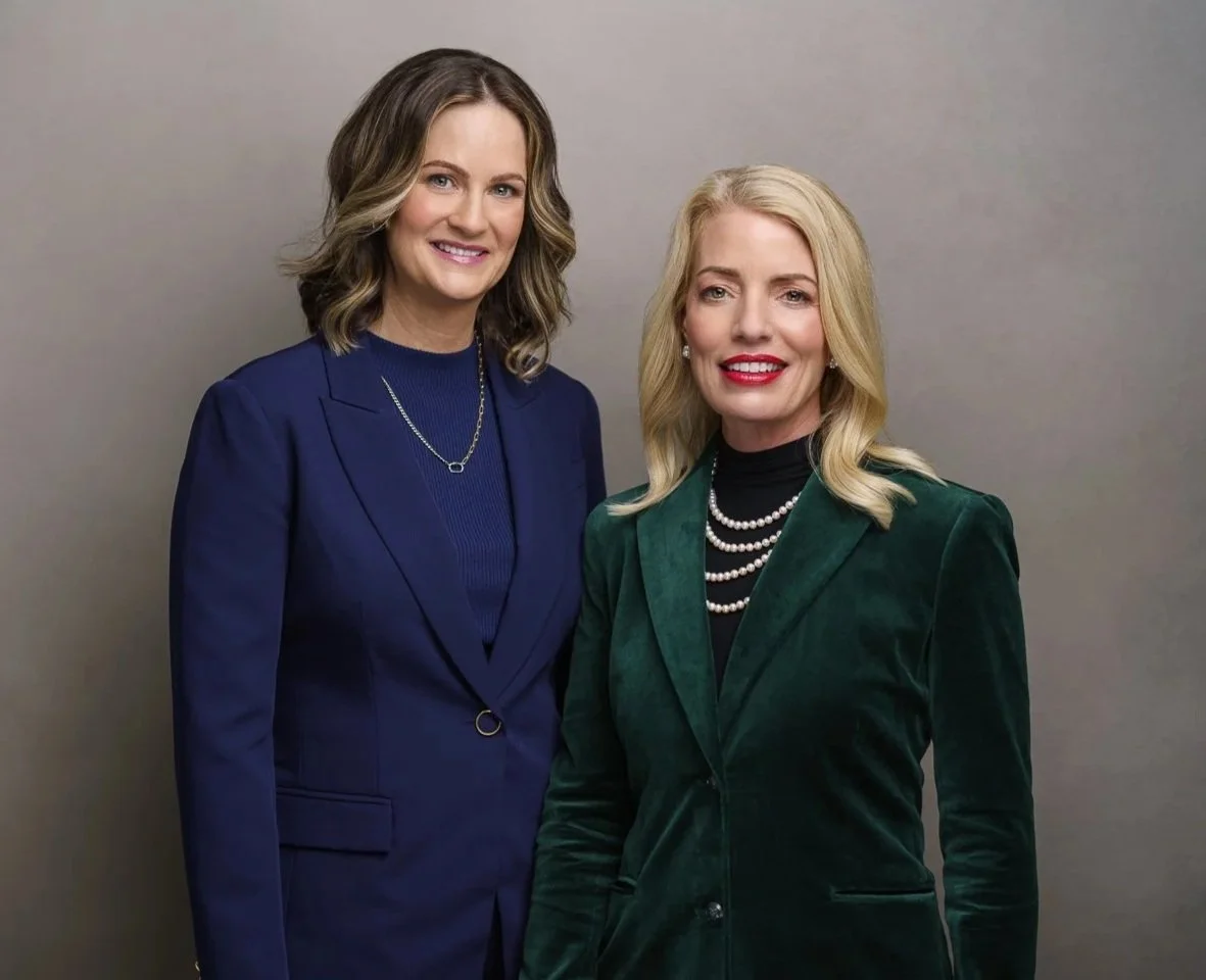 Two women dressed professionally standing side by side in front of a plain gray background, smiling at the camera.