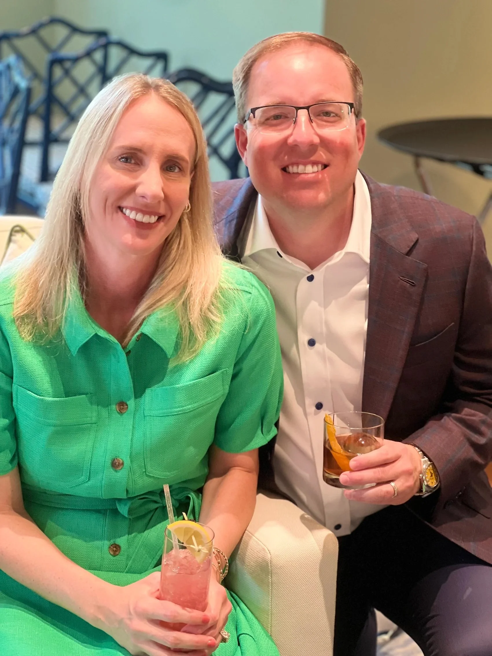 A woman in a bright green dress and a man in a suit with glasses sitting together at a table, smiling at the camera, each holding a drink