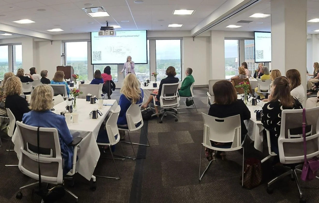 Laura K. Sawyier is giving a presentation to a seated audience in a conference room with large windows showing a cityscape. 