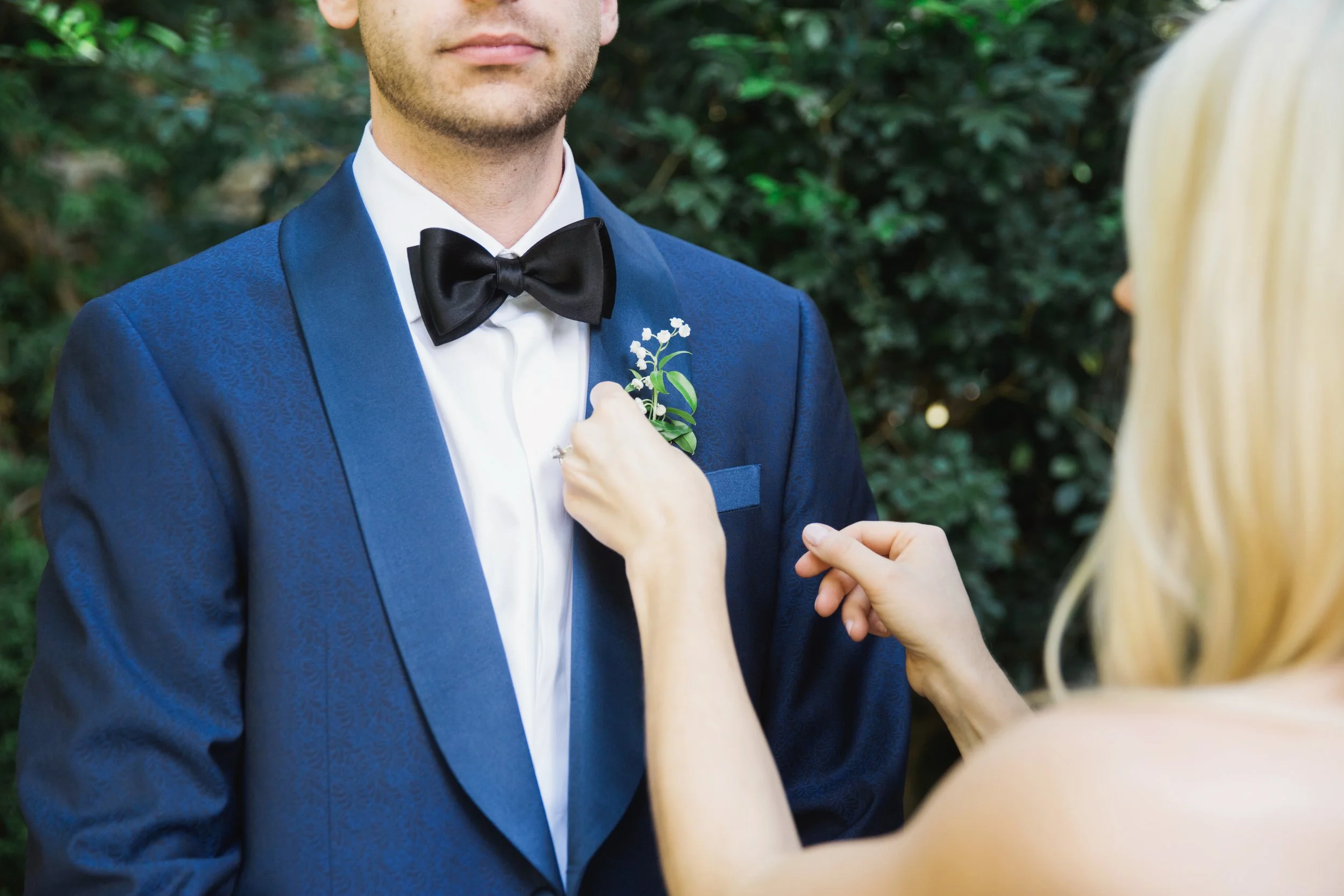 A woman pins a boutonniere onto a man's blue suit jacket during a wedding or formal event, set outdoors with greenery in the background.