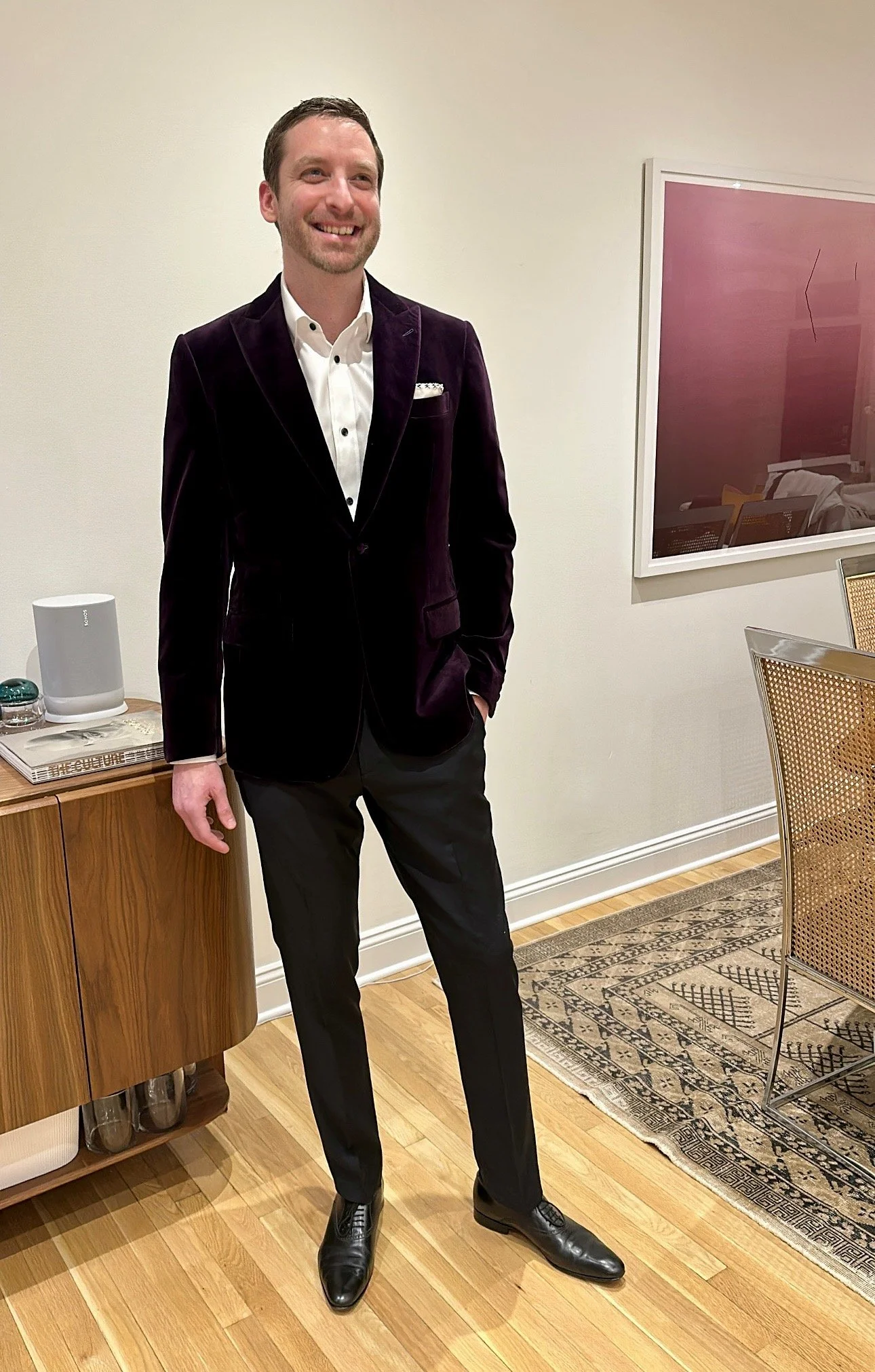 A man dressed in a dark velvet blazer, white shirt, and black trousers stands in a room with wood flooring, smiling.