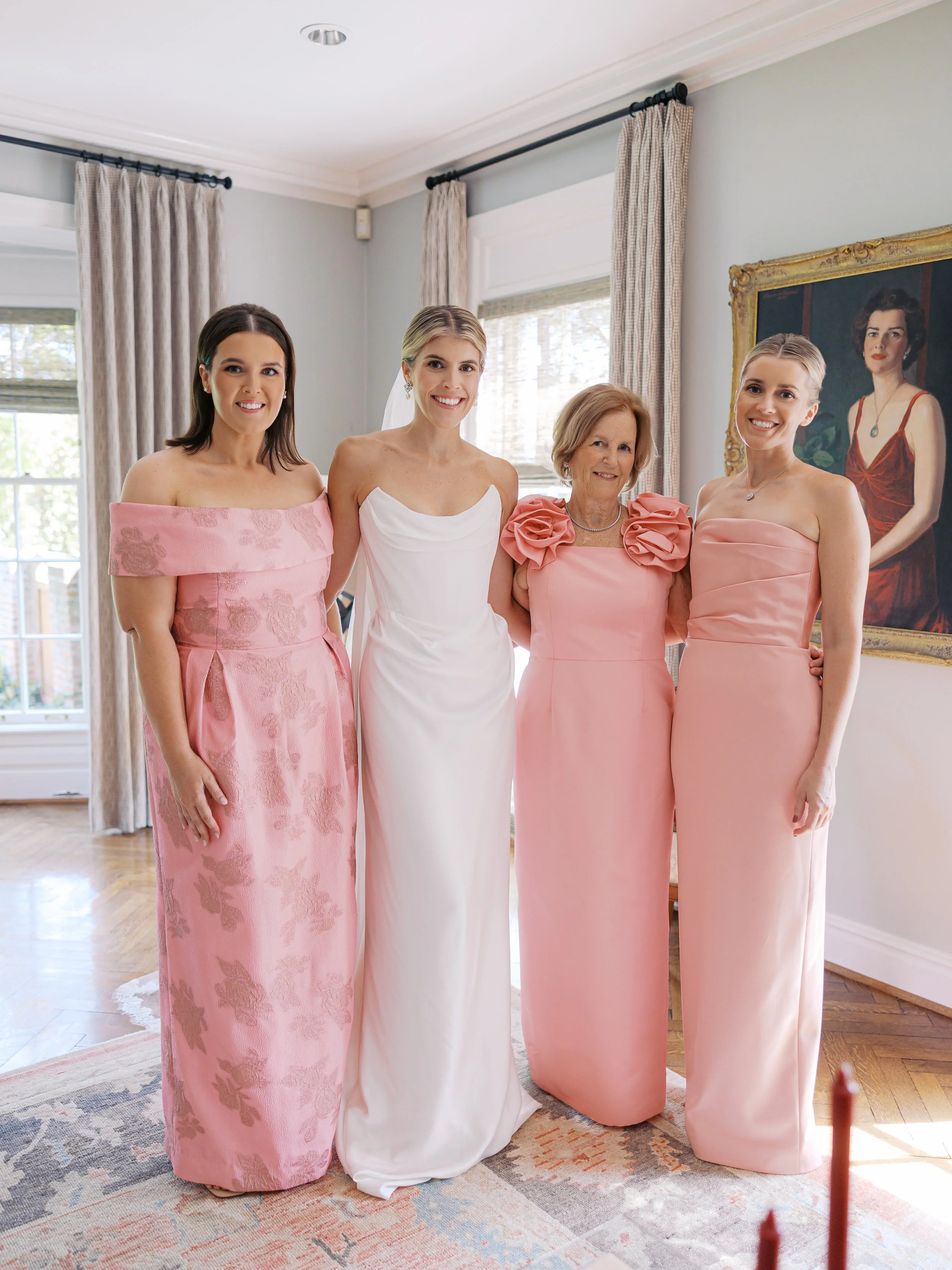 Four women, including a bride in a white wedding gown, standing together in a well-lit room with beige curtains and a large portrait painting on the wall.