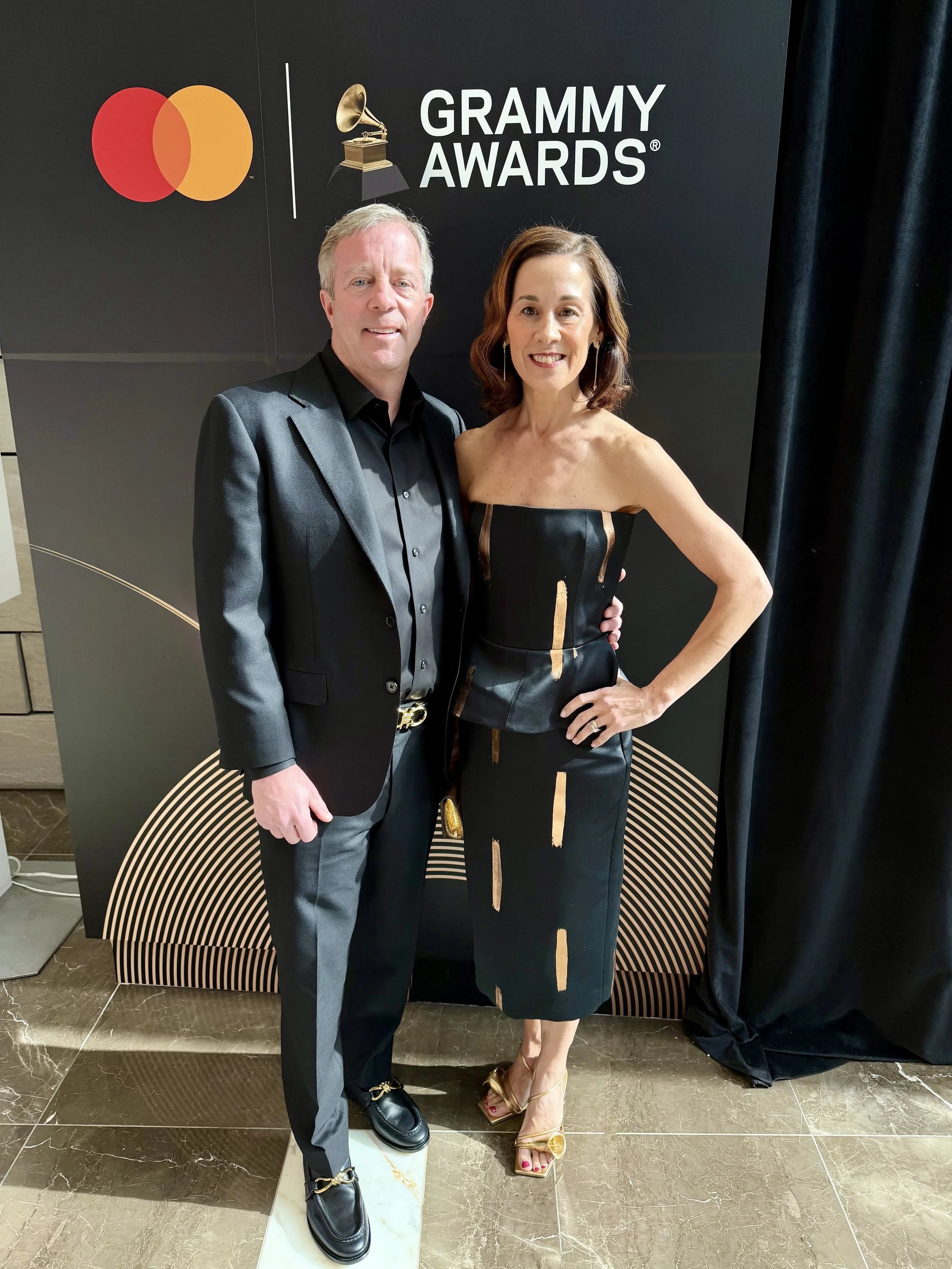 A man and a woman posing together on the red carpet at the Grammy Awards, with a black backdrop showing the Grammy Awards logo and the Mastercard logo.
