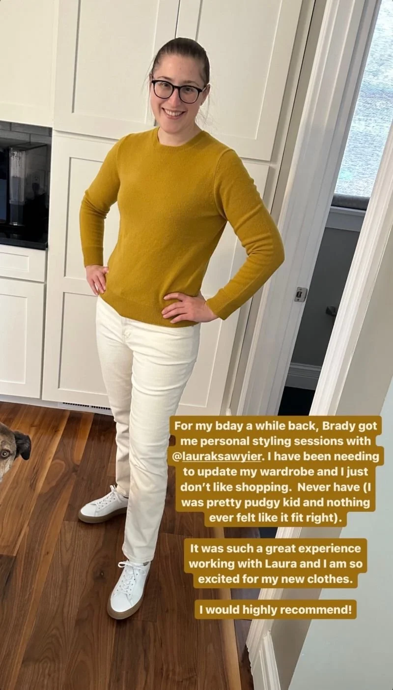 A woman with glasses and a ponytail, wearing a mustard yellow sweater and white pants, standing in a kitchen with her hands on her hips, smiling at the camera.