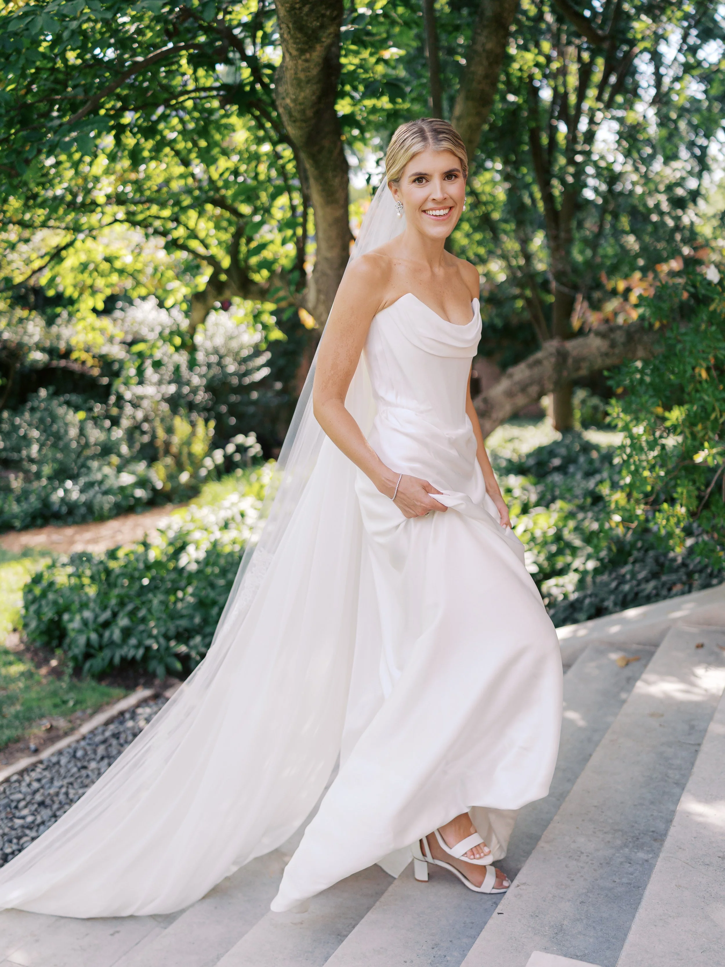 A bride in a white wedding gown with a long train, standing outdoors on a stone staircase surrounded by green trees and bushes, smiling and looking at the camera.