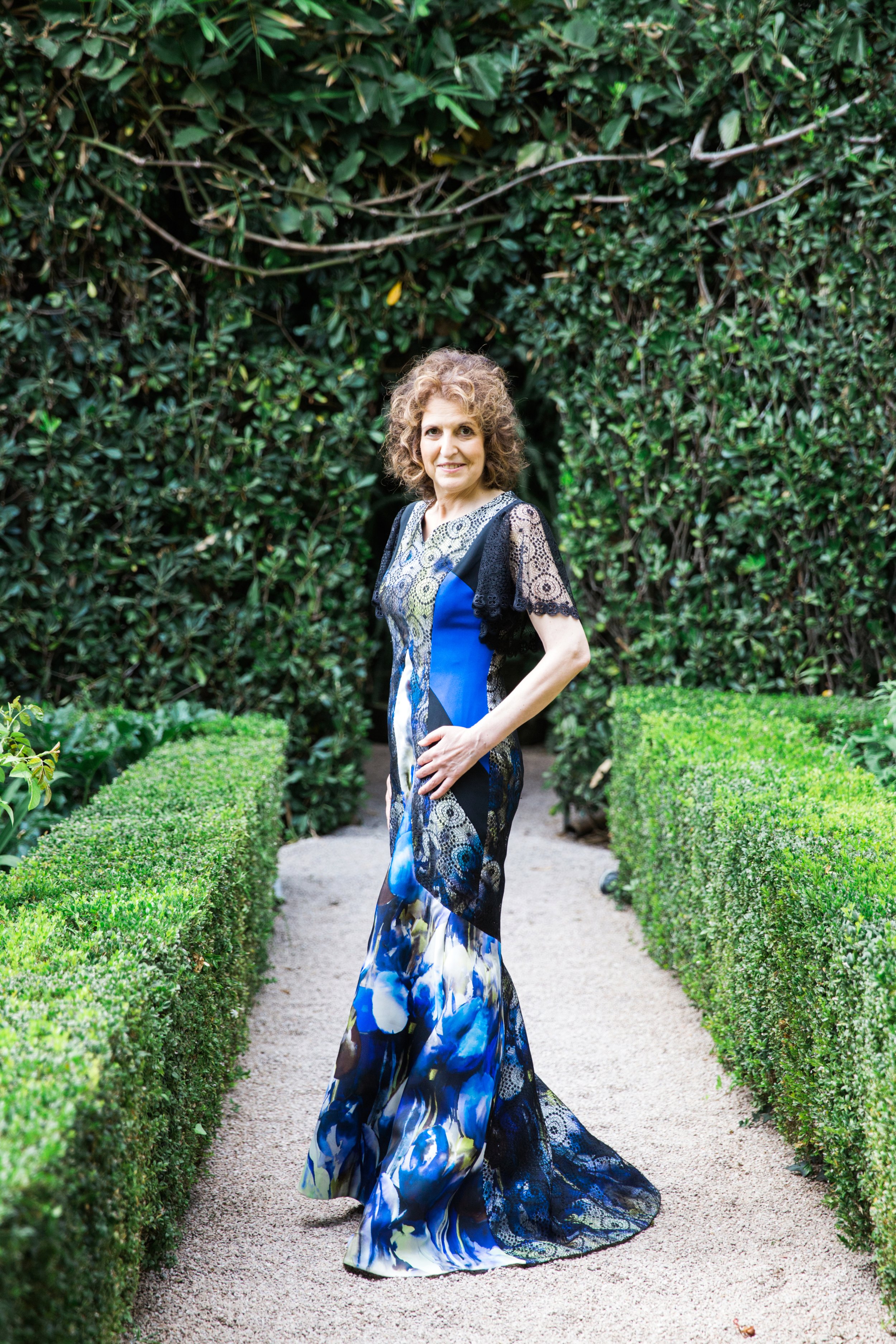 A woman with curly hair standing on a gravel path, surrounded by green hedges and a lush backdrop of trees and bushes.