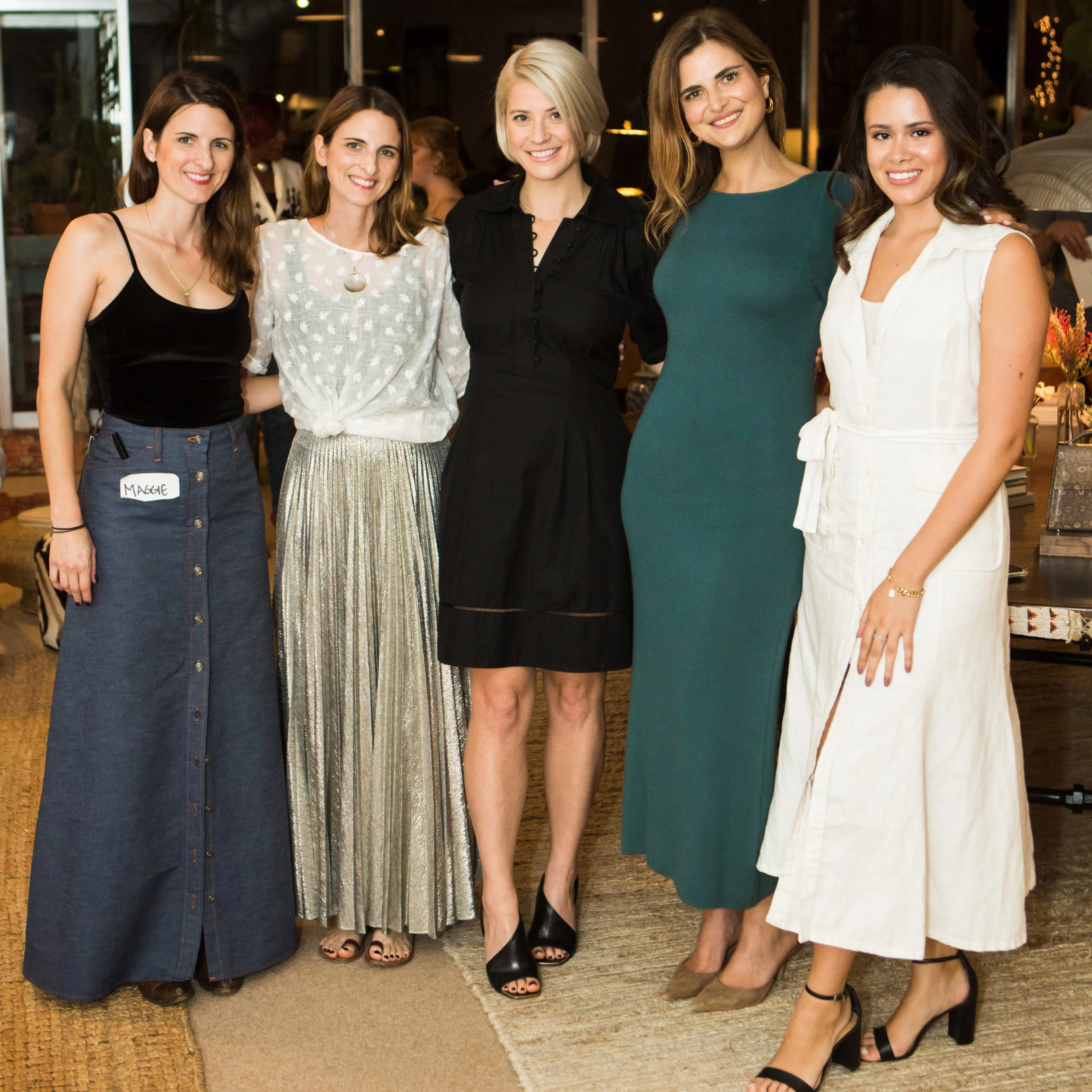 Five women standing together indoors, smiling at the camera. They are dressed in stylish outfits and are in a warmly lit setting.