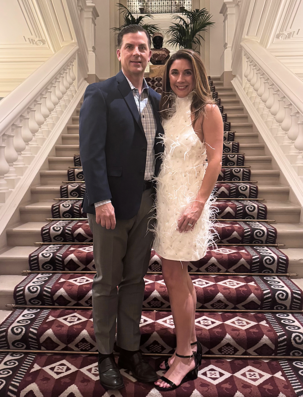 A man and woman stand together on a staircase decorated with a patterned rug, with potted plants and sculptures visible in the background, dressed formally for an event.
